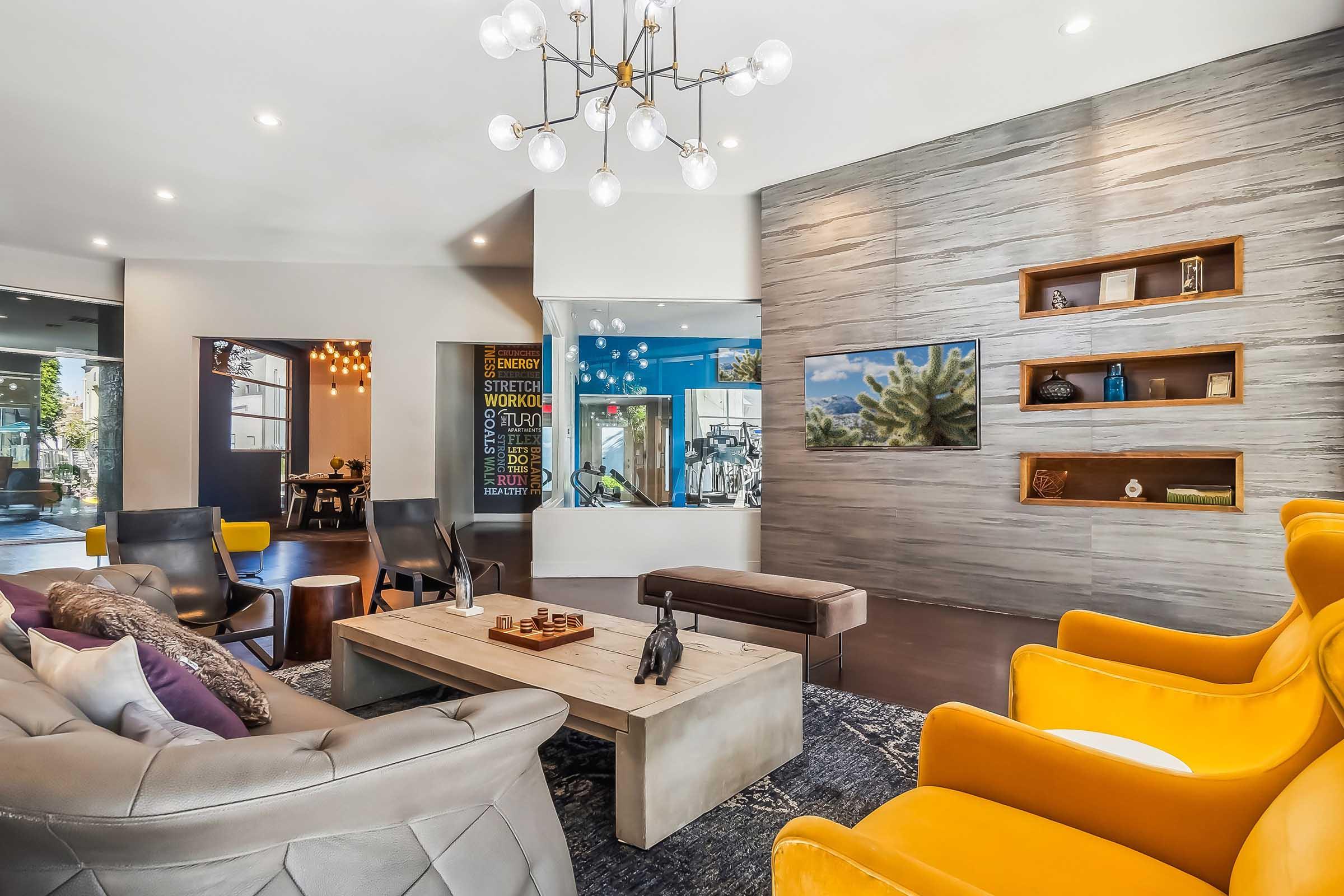 A contemporary lounge area featuring gray and yellow armchairs, a wooden coffee table, and decorative elements. A large window allows natural light, while a wall-mounted TV displays an artwork. Modern lighting fixtures add a stylish touch to the inviting space.