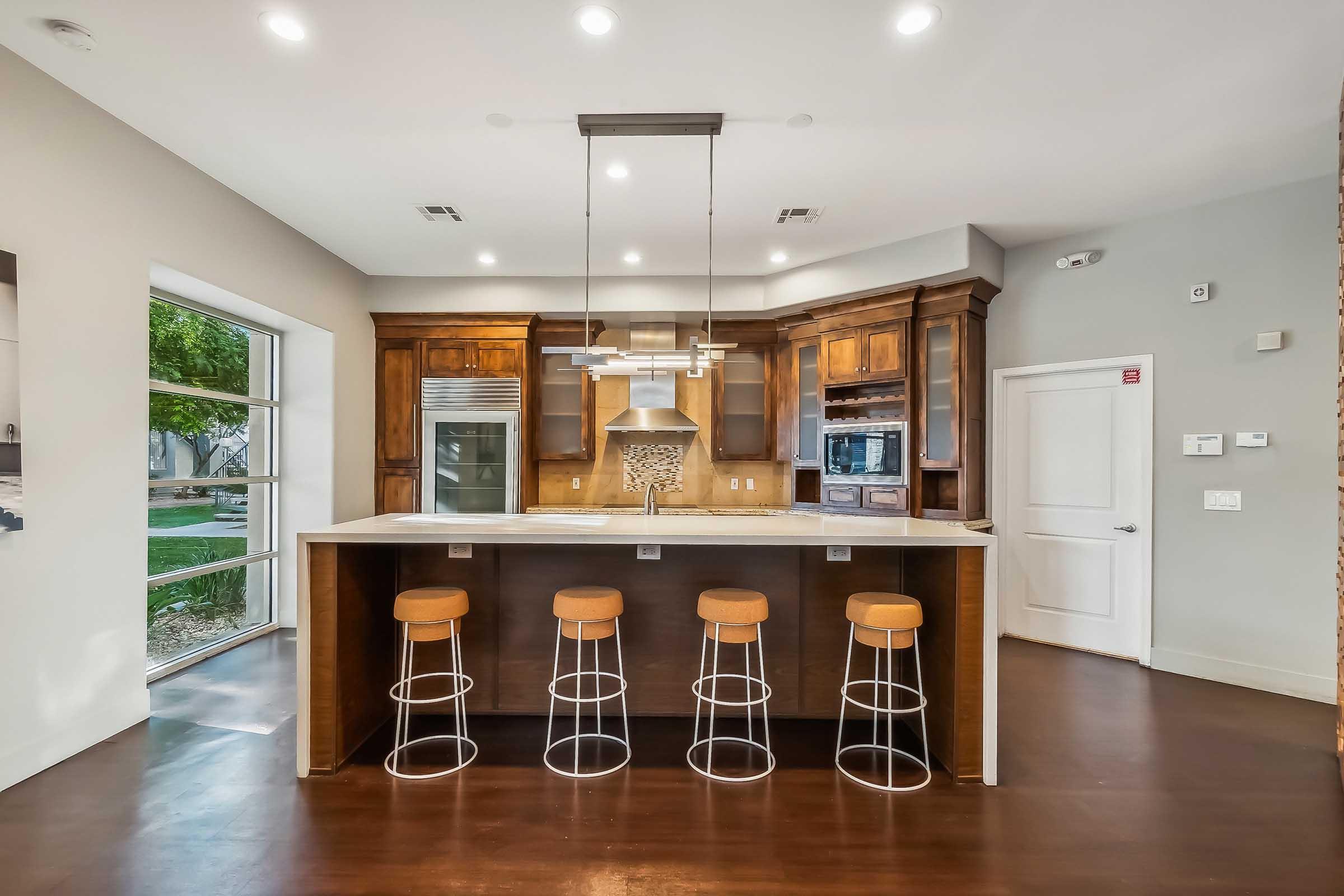 A modern kitchen featuring wooden cabinetry, a central island with four stools, and stainless steel appliances. The area is well-lit with recessed lighting and a window allowing natural light to enter, showcasing a clean and contemporary design.