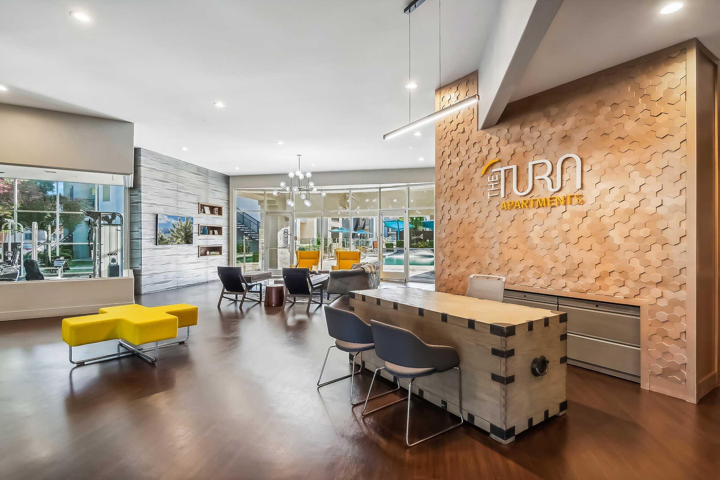 Modern apartment lobby featuring a welcoming reception desk, stylish seating areas with yellow accents, and large windows allowing natural light. The wall showcases a textured design with the name "The Turn Apartments" prominently displayed. A fitness area is visible through the glass, enhancing a vibrant communal atmosphere.