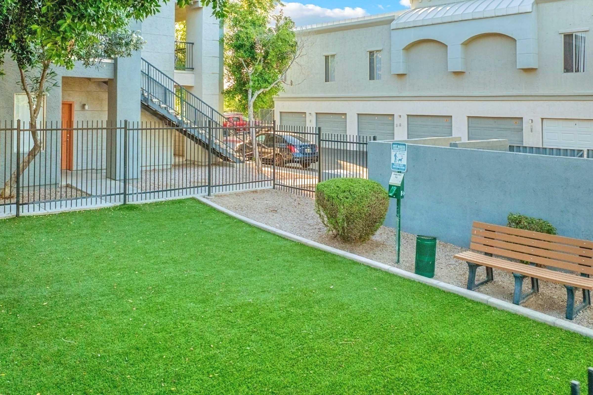 A well-maintained outdoor area featuring a green lawn, a bench, and a small bush. In the background, there's an apartment building with a staircase and several parking spaces. The scene is bright and inviting, with trees providing some shade.
