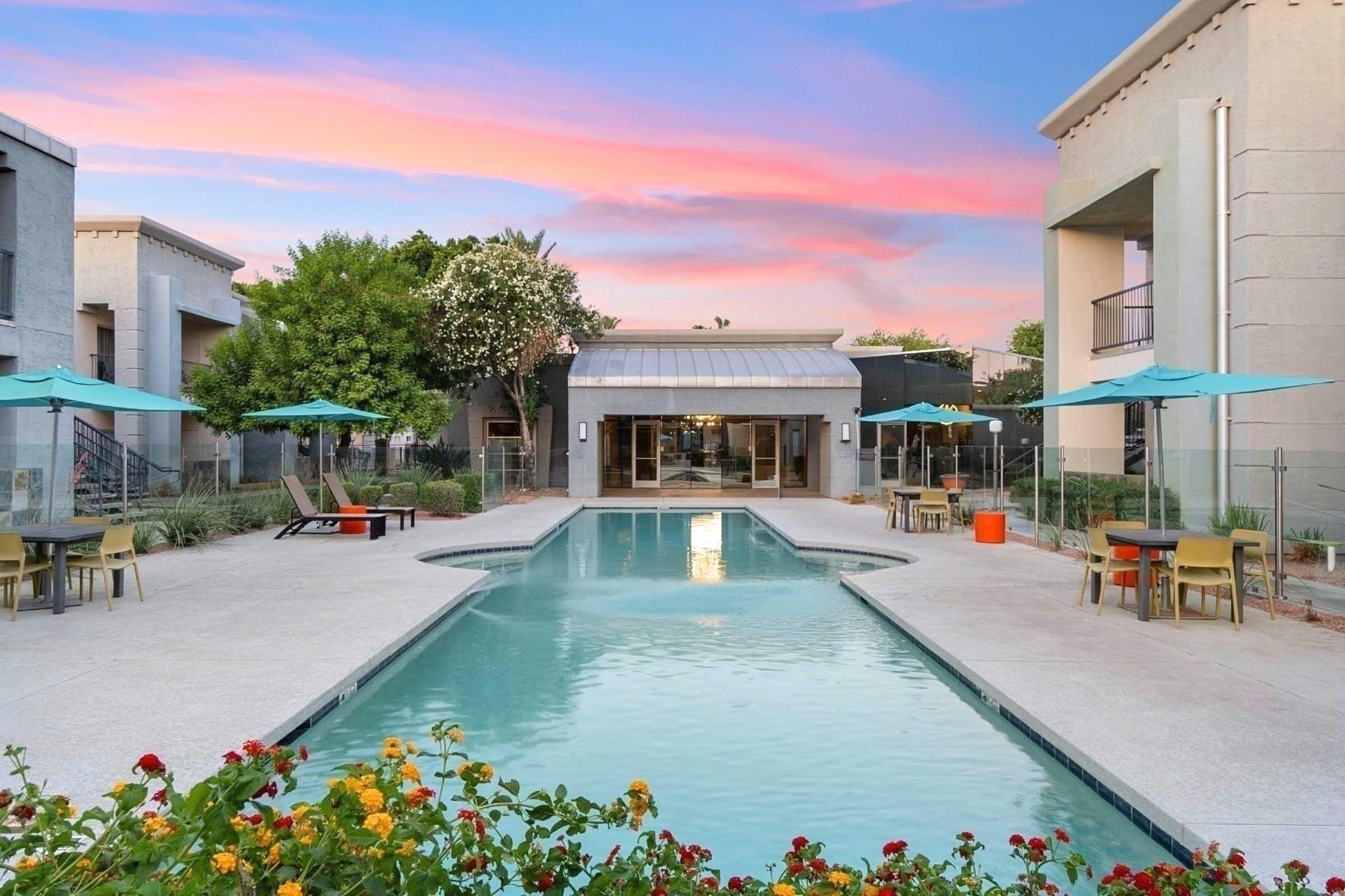 A serene outdoor pool area surrounded by colorful flowers and green landscaping. Umbrella-covered tables and chairs are set near the pool, with a modern building in the background. The sky is painted in soft pink and blue hues, creating a tranquil atmosphere.