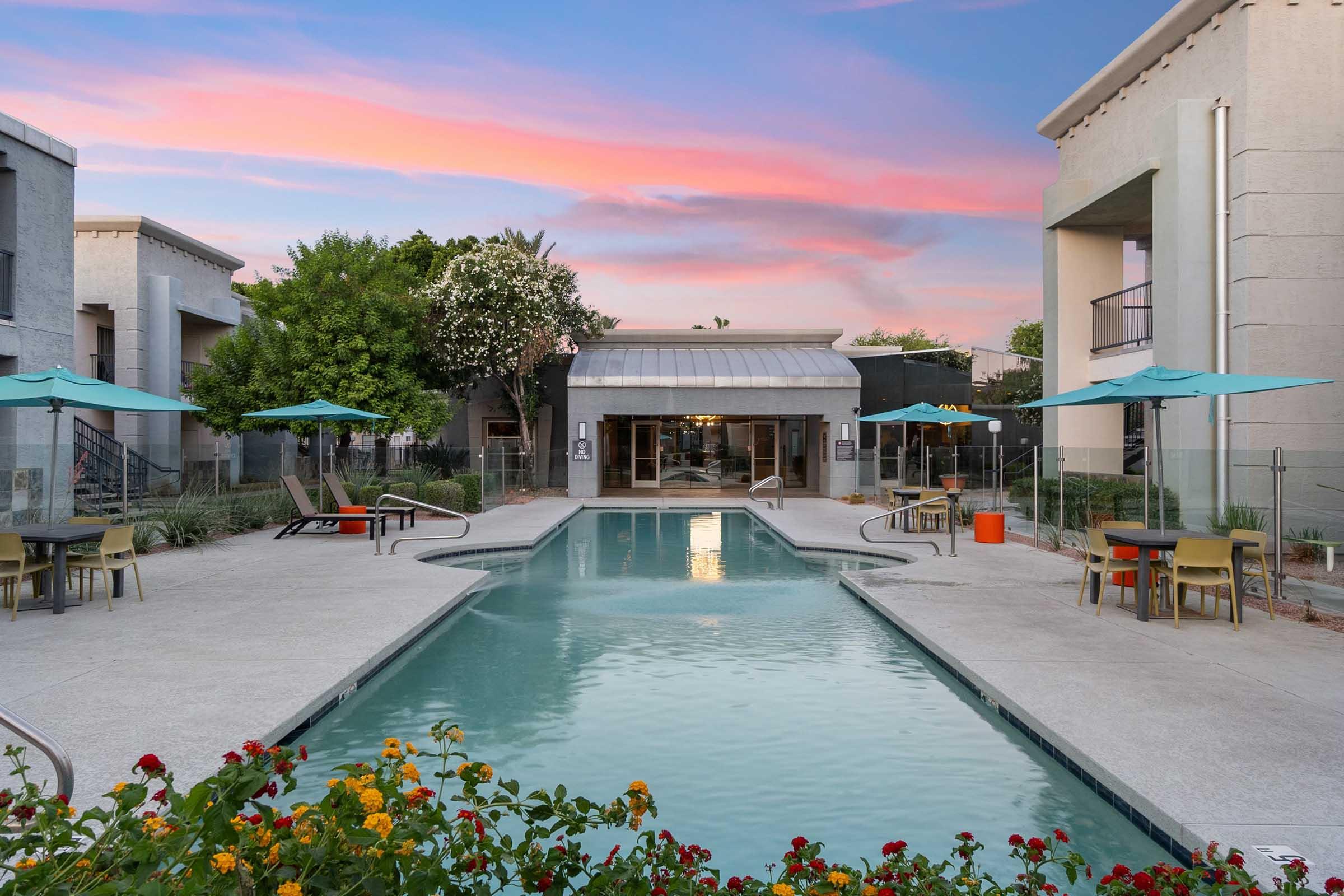 A serene outdoor pool surrounded by modern buildings, with vibrant flowers in the foreground. Umbrella-covered tables and lounge chairs are arranged around the pool area. The sky displays soft pink and blue hues, suggesting either sunrise or sunset, contributing to a tranquil atmosphere.