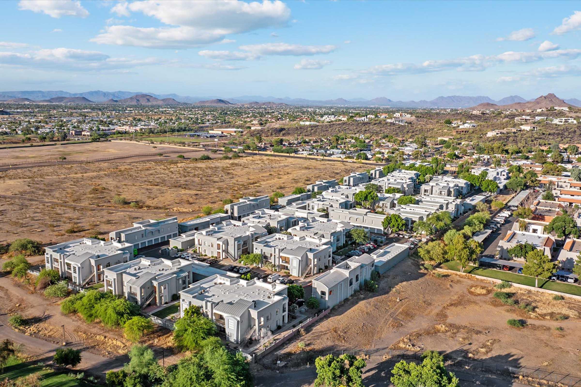 Aerial view of a residential area featuring several modern apartment buildings surrounded by green trees and open land. In the background, rolling hills and mountains are visible under a clear blue sky with scattered clouds. The scene showcases a blend of urban development and natural landscape.
