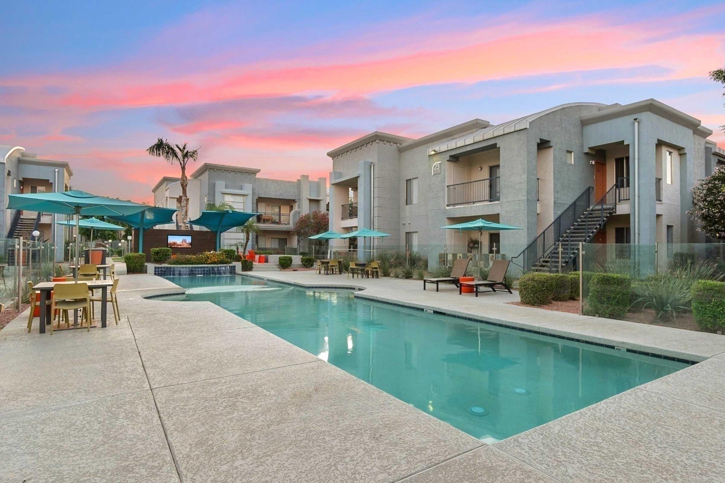 A serene apartment complex featuring a sparkling swimming pool surrounded by lounge chairs and tables. Palm trees and umbrellas provide shade, with elegant buildings in the background under a colorful sunset sky. The ambiance is inviting and perfect for relaxation.