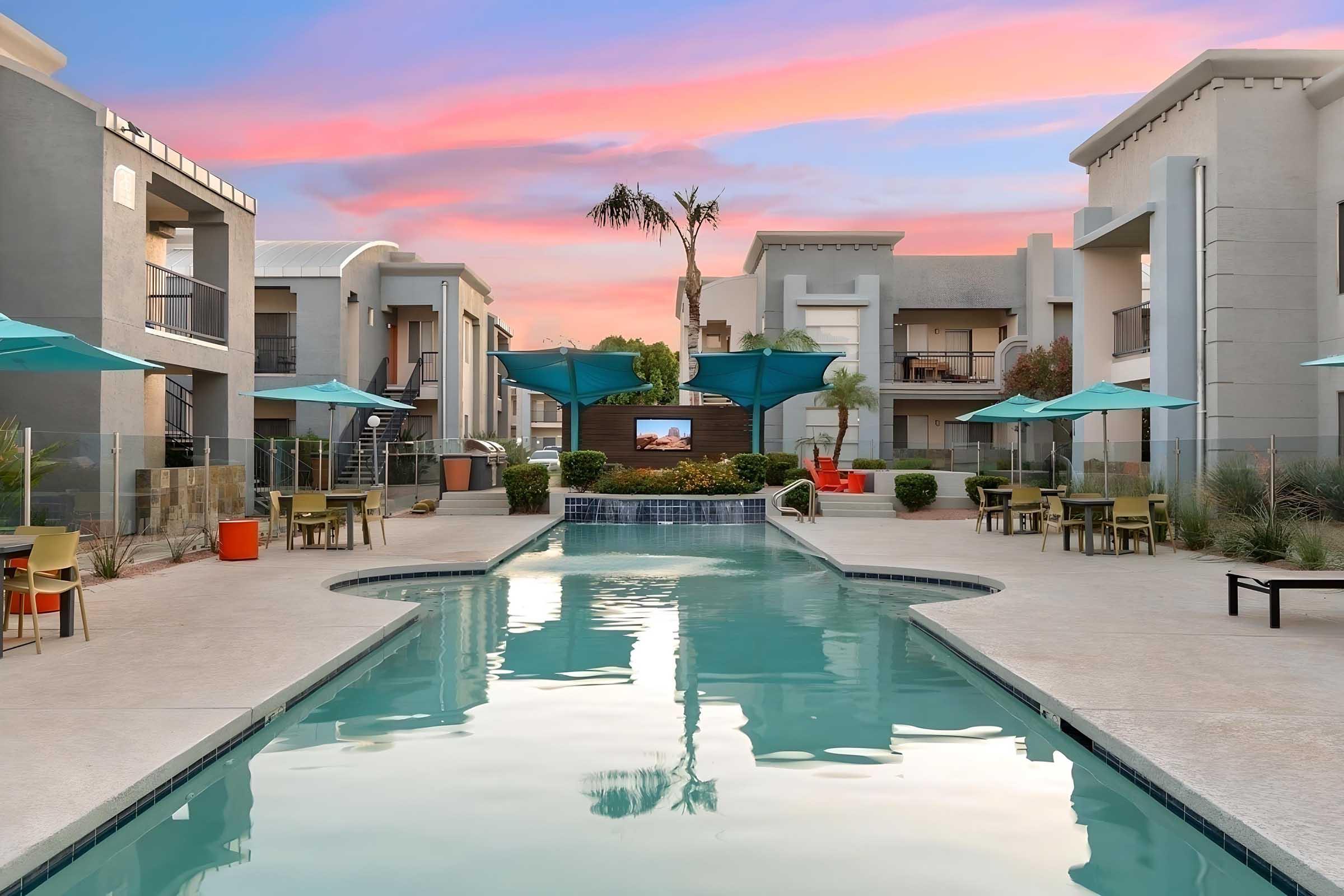 A serene swimming pool surrounded by modern apartment buildings, featuring lounge chairs and umbrellas. The sky is painted with pink and orange hues during twilight, creating a tranquil atmosphere. Lush landscaping and a small waterfall enhance the relaxing outdoor setting.