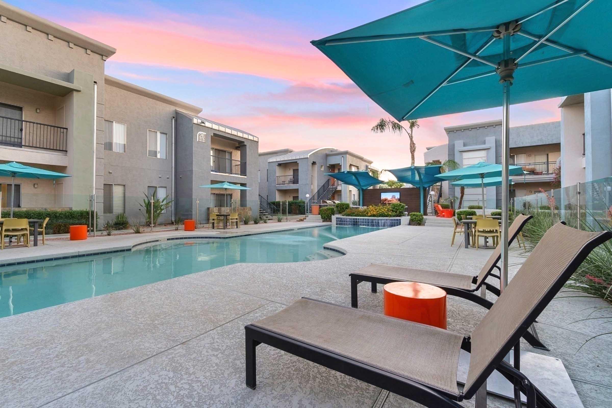 A modern apartment complex with a swimming pool surrounded by lounge chairs and turquoise umbrellas. The sky is painted in pink hues during sunset, and there are potted plants and outdoor furniture, creating a relaxed and inviting atmosphere.