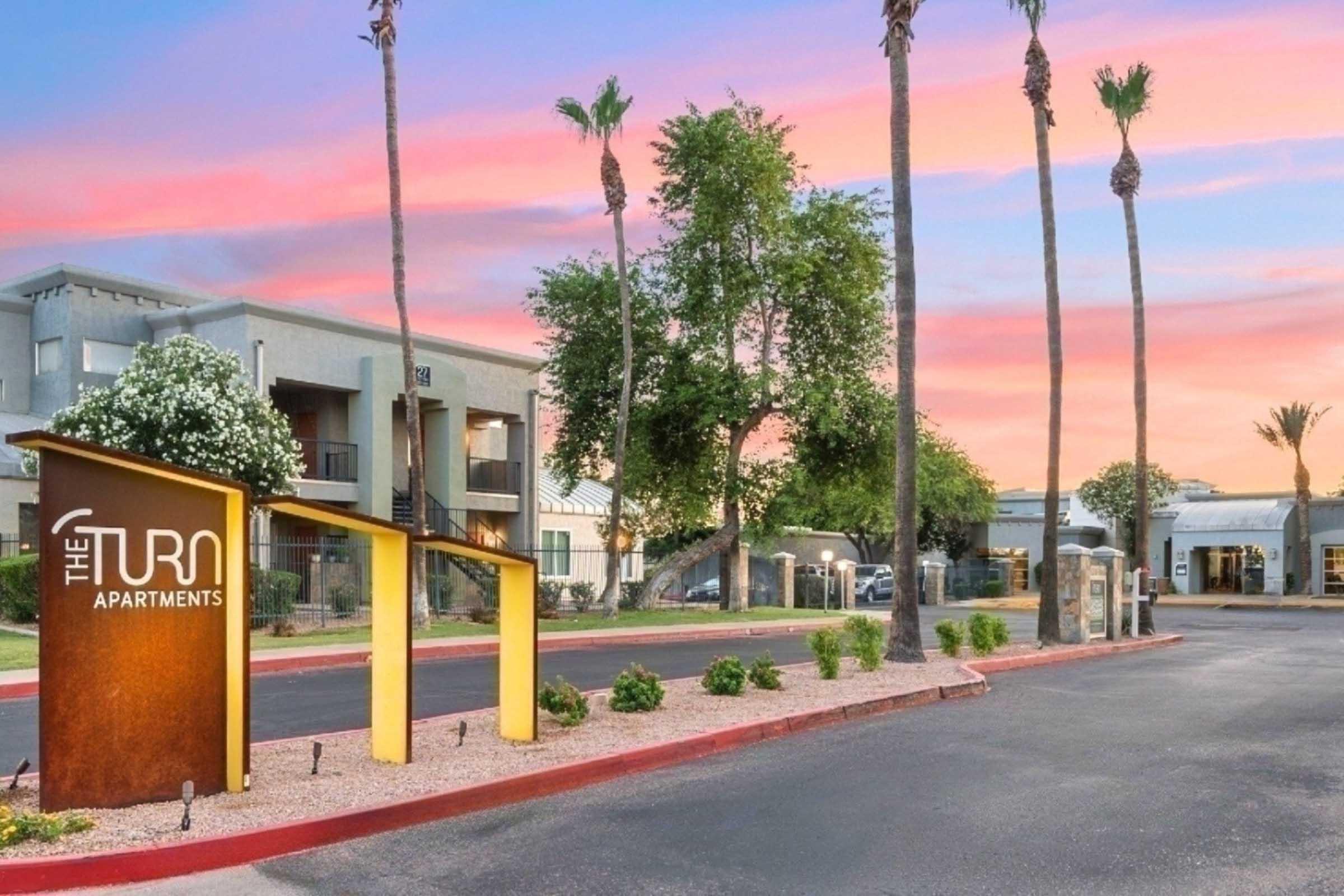 Exterior view of The Turn Apartments featuring modern architecture, palm trees, colorful sunset skies, and a welcoming entrance sign. The surrounding area is landscaped with gravel and greenery, creating an inviting atmosphere for potential residents.