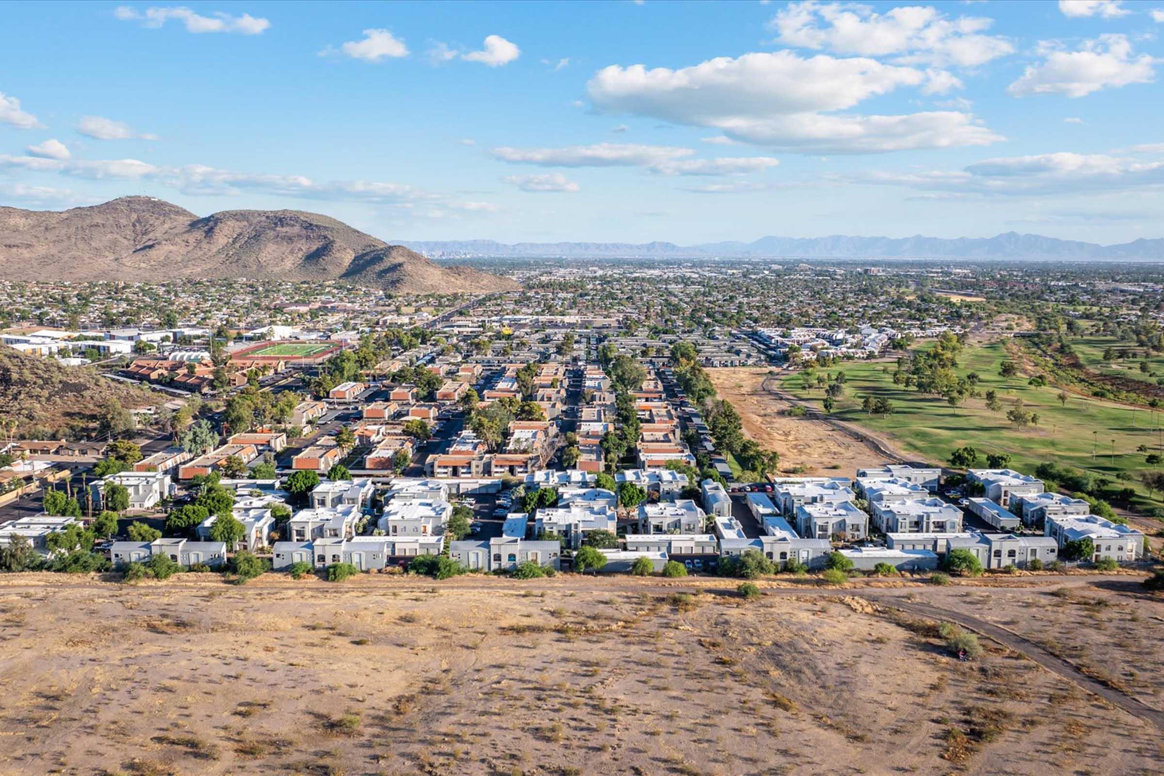 Aerial view of a suburban area with clusters of houses interspersed with greenery. In the background, rolling hills and mountains are visible under a partly cloudy blue sky. The landscape features residential neighborhoods, open spaces, and some golf courses.