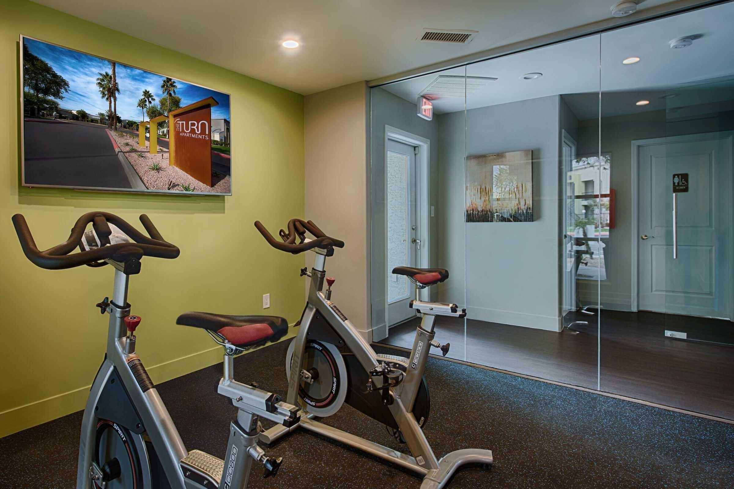 A fitness room featuring two stationary bikes, a large wall-mounted television displaying a scenic view, and a glass wall that leads to a restroom. The walls are painted in a soothing green color, and the floor has a textured black surface. Natural lighting comes from a nearby window.