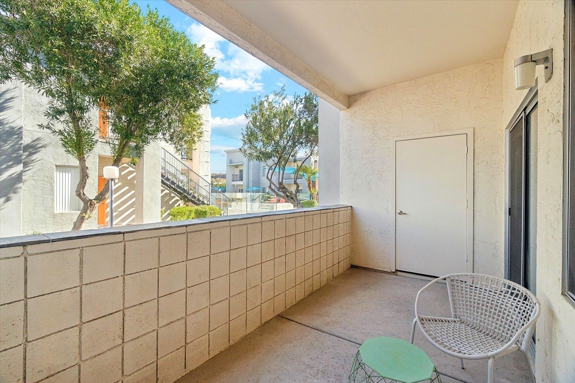 A spacious balcony with a white wicker chair and a small green side table. The balcony is surrounded by light-colored walls and overlooks a courtyard with trees and nearby buildings under a blue sky. Sunlight casts soft shadows on the floor.