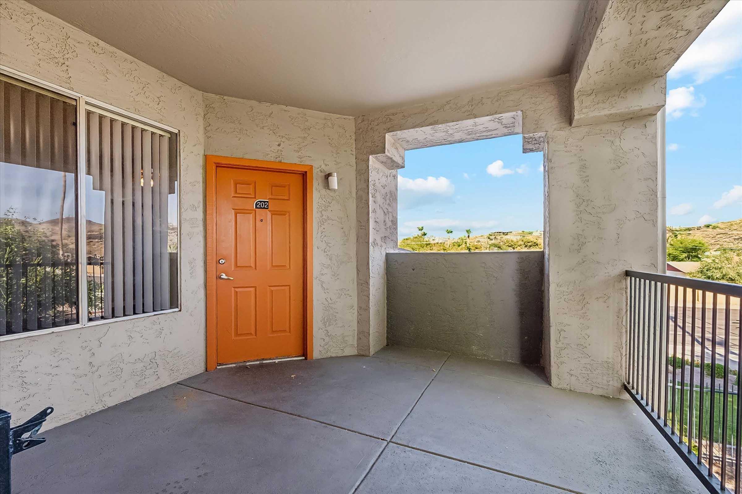 View of a balcony area featuring an orange door leading into a unit. The walls are textured in a light color, with large windows covered by vertical blinds. There is a railing and an open view showing greenery and distant hills in the background under a blue sky with some clouds.