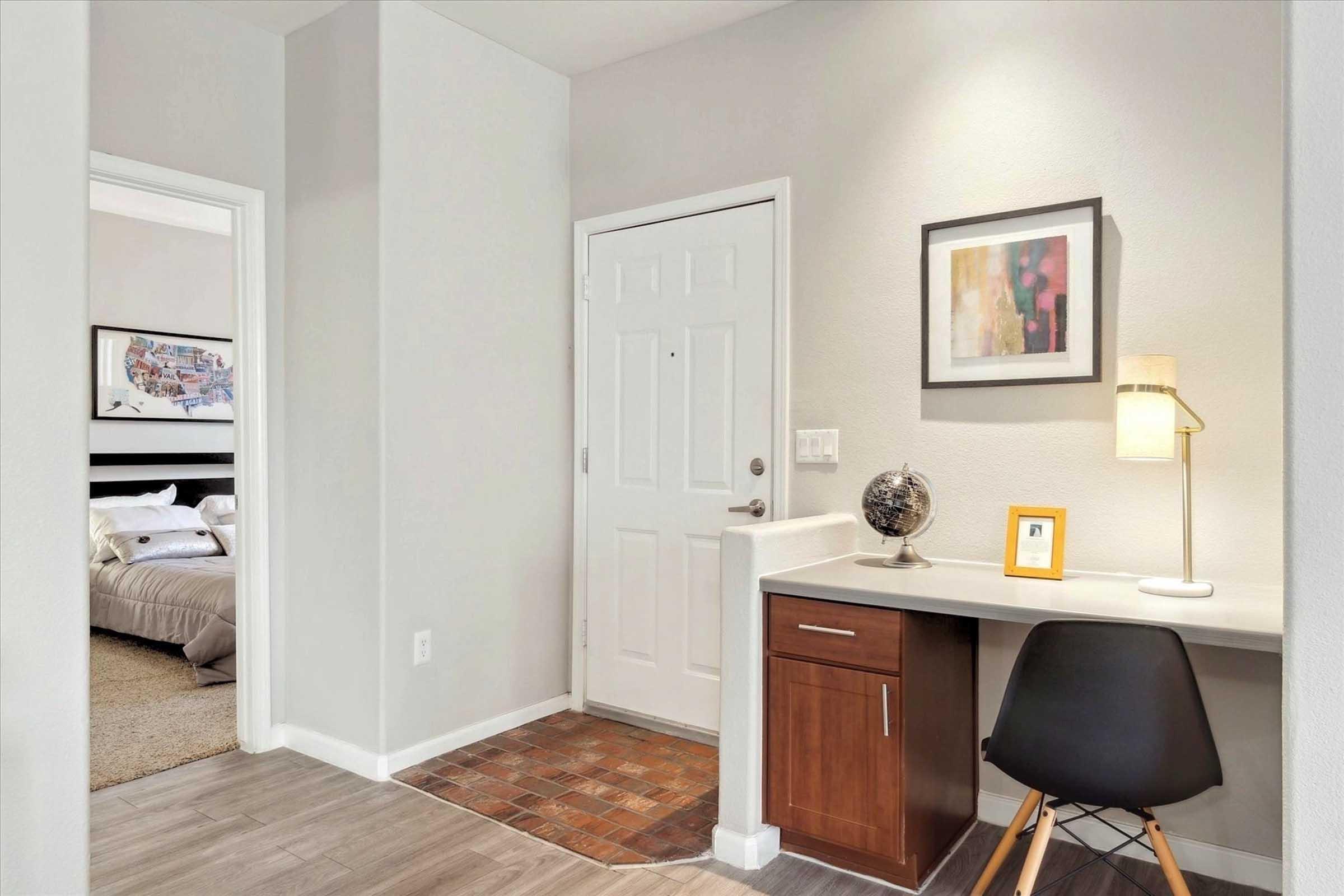 A stylish entryway featuring a small desk with a lamp and globe, a door leading to a bedroom, and a decorative framed picture on the wall. The area is well-lit and has neutral colors with a textured floor and a cozy atmosphere.