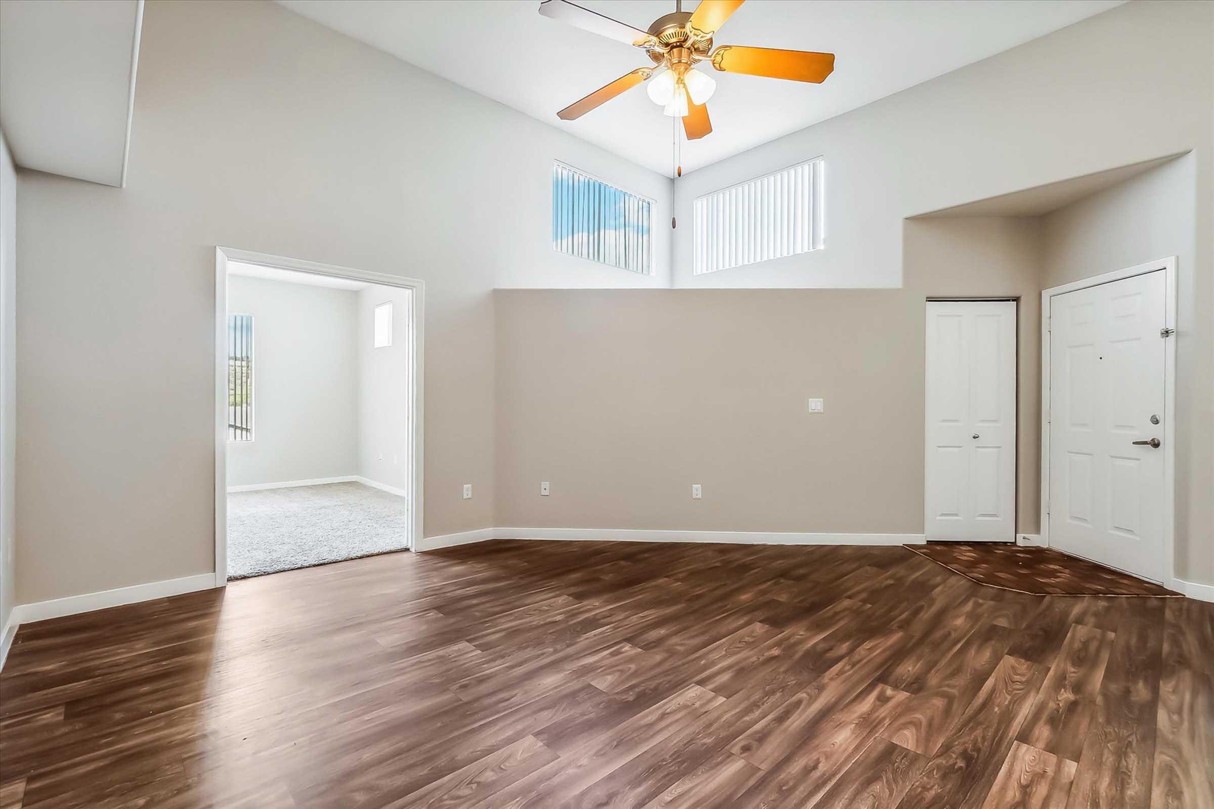 A spacious interior view of a modern living area featuring a ceiling fan, large windows for natural light, and a rich wooden floor. An entrance door is visible, along with an open doorway leading to another room. The walls are painted in a light color, creating a bright and airy atmosphere.