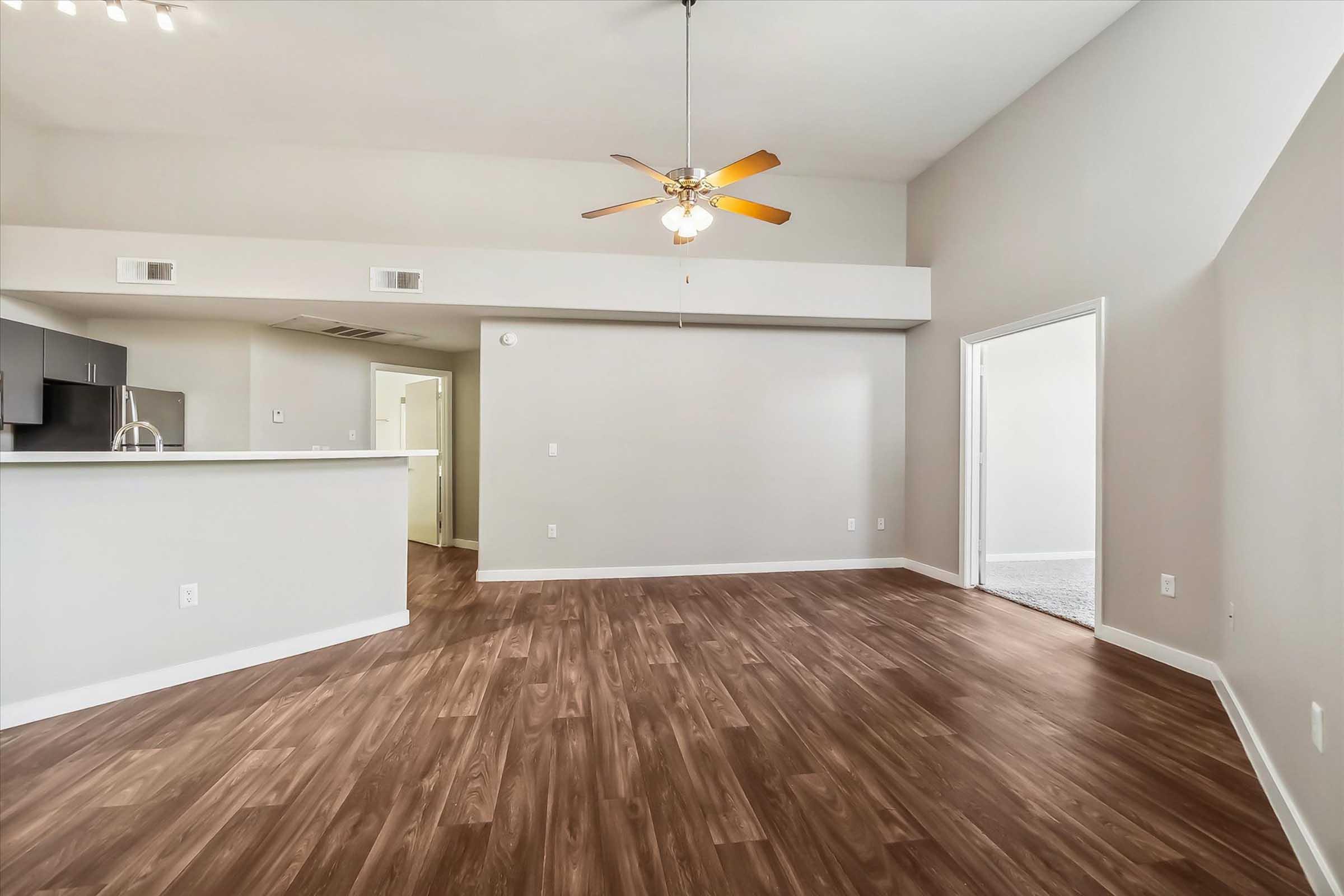 A spacious living area featuring light-colored walls, a ceiling fan with wooden blades, and hardwood flooring. There is an open kitchen area with dark cabinets visible, along with an open doorway leading to another room. Natural light from windows enhances the bright atmosphere of the space.