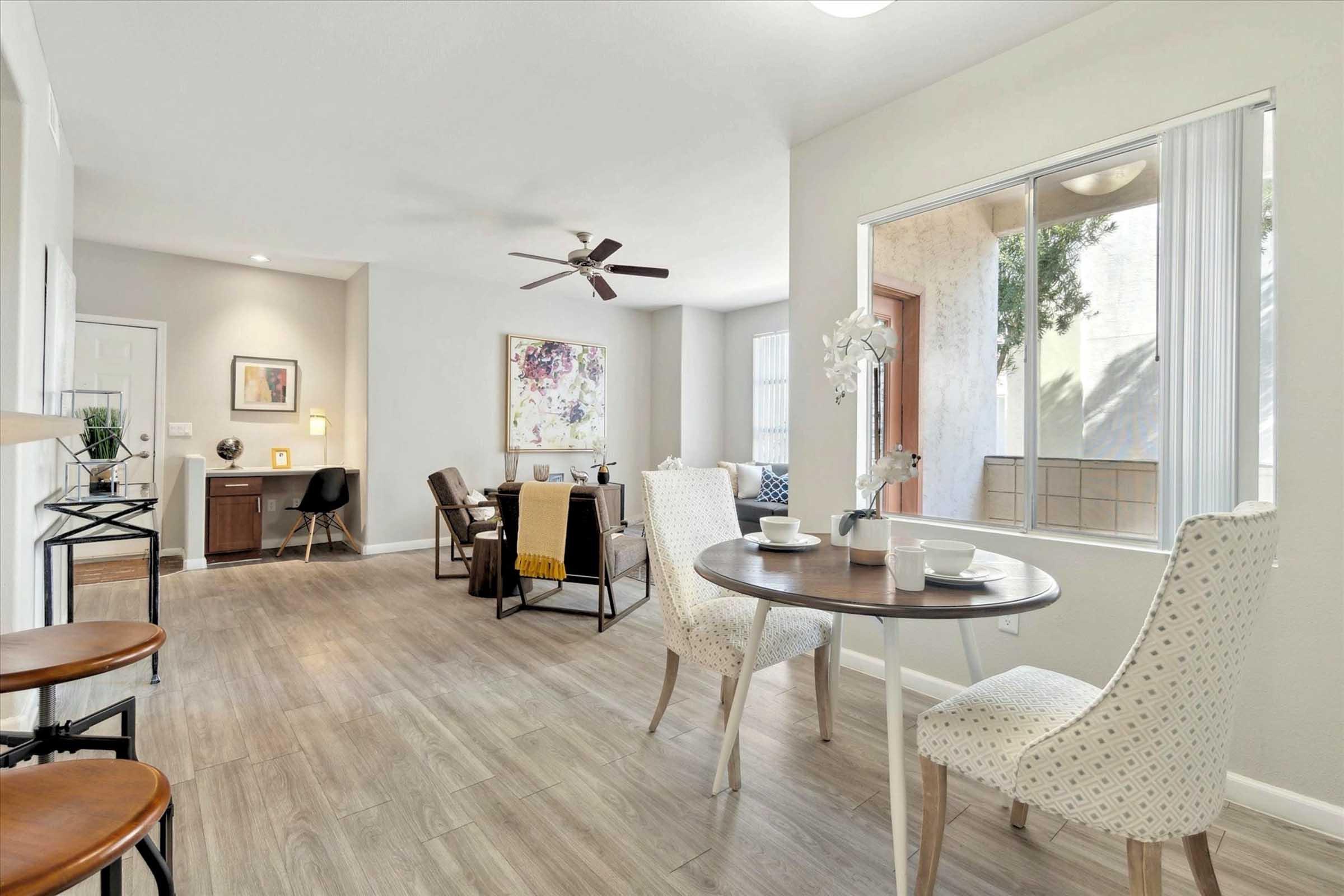 A stylish, modern living space featuring a dining area with a round table and two chairs, adorned with tableware. The room has a cozy seating area with a desk, artwork on the walls, and large windows allowing natural light to flow in. Light-colored walls and wooden flooring create a warm atmosphere.