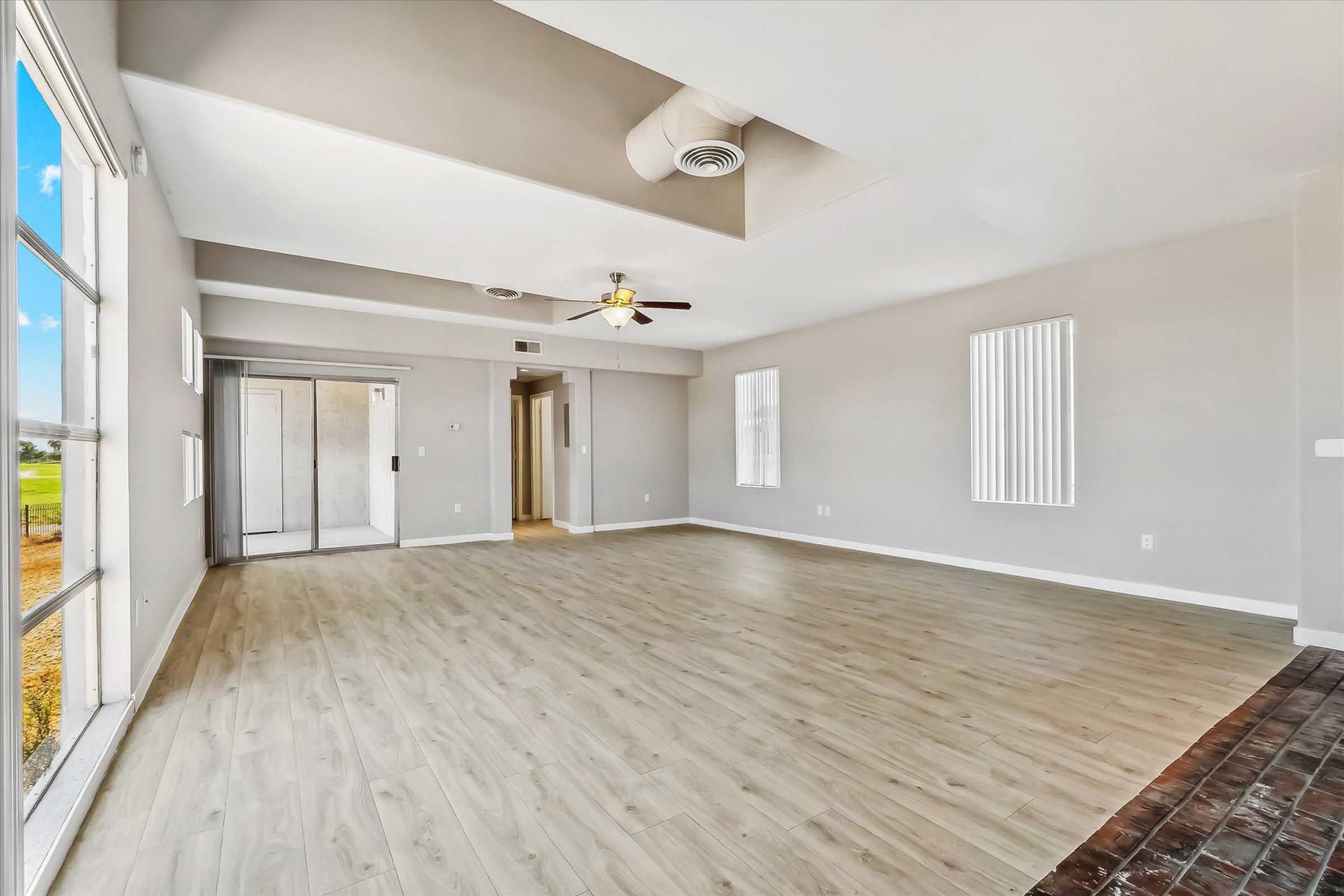 Spacious interior of a room with light grey walls and wooden flooring. It features large windows allowing natural light, a ceiling fan, and sliding closet doors. The room has a modern, open layout suitable for various furniture arrangements.