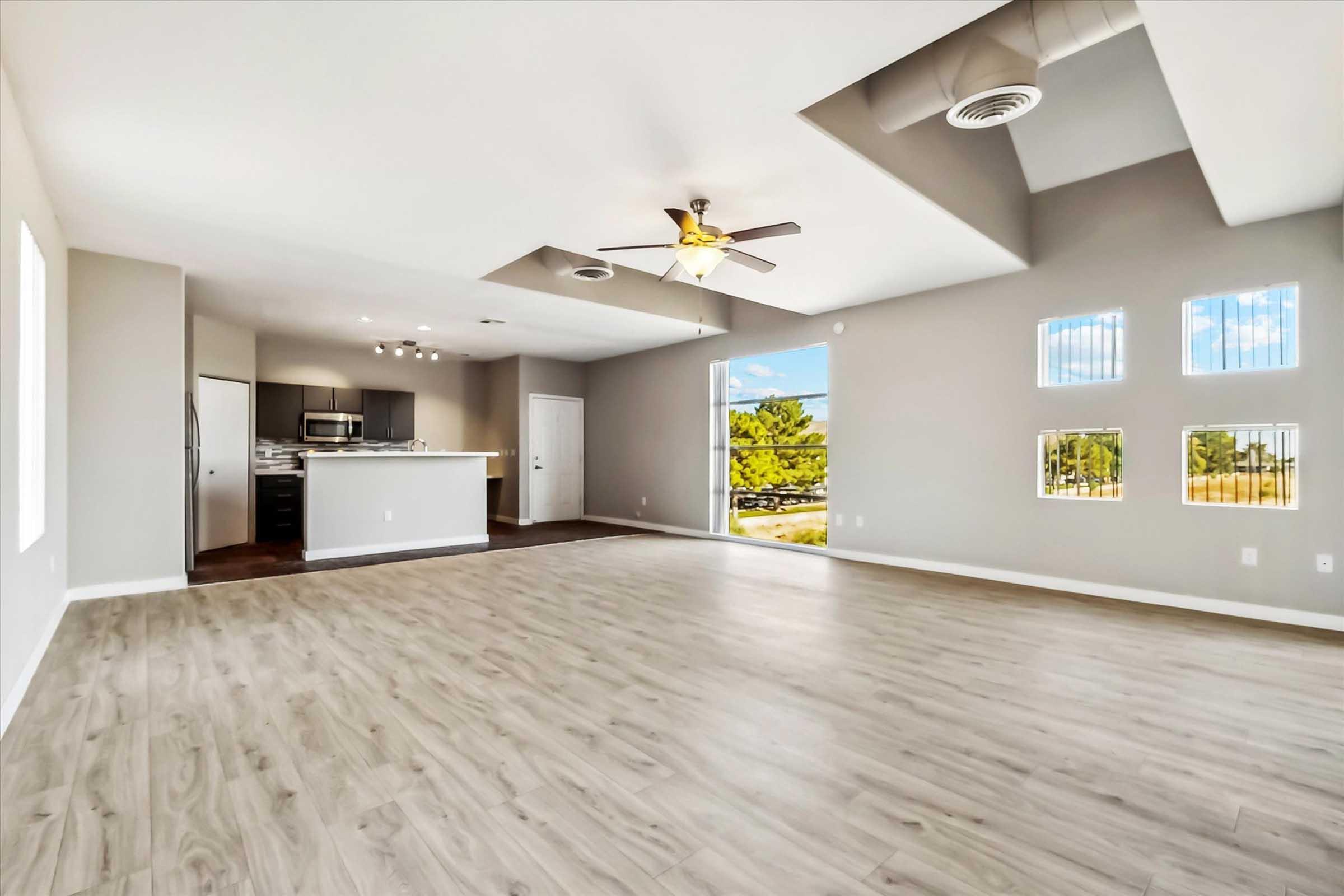 Spacious modern living room with light hardwood flooring, a ceiling fan, and large windows. The kitchen is partially visible in the background, featuring dark cabinetry and stainless steel appliances. The walls are painted in a neutral color, creating a bright and airy atmosphere.