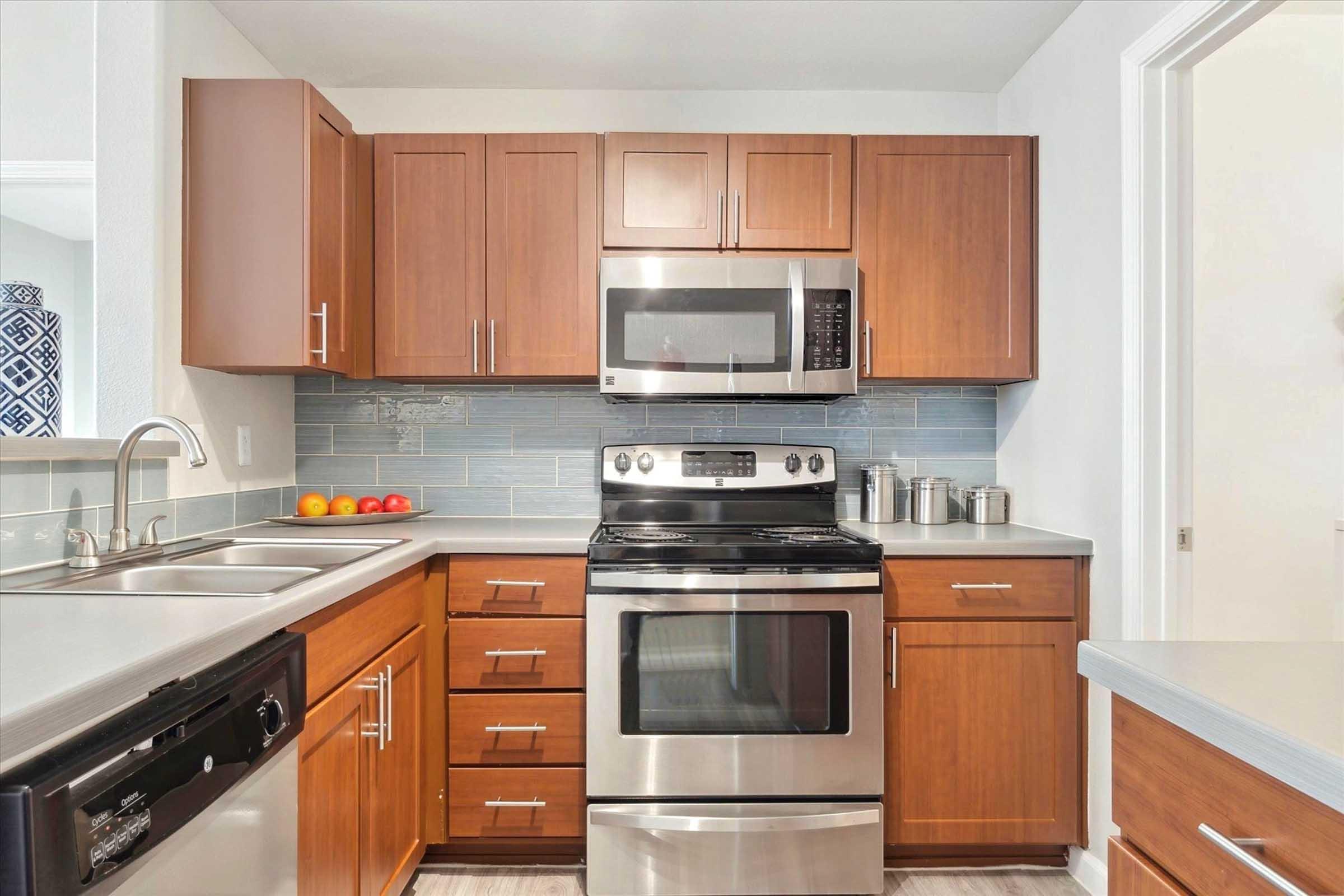 Modern kitchen featuring wooden cabinetry, stainless steel appliances including a microwave and stove, and a sink. Countertop space is visible, adorned with a bowl of apples. The backsplash is a light blue tile, adding a touch of color to the overall design.