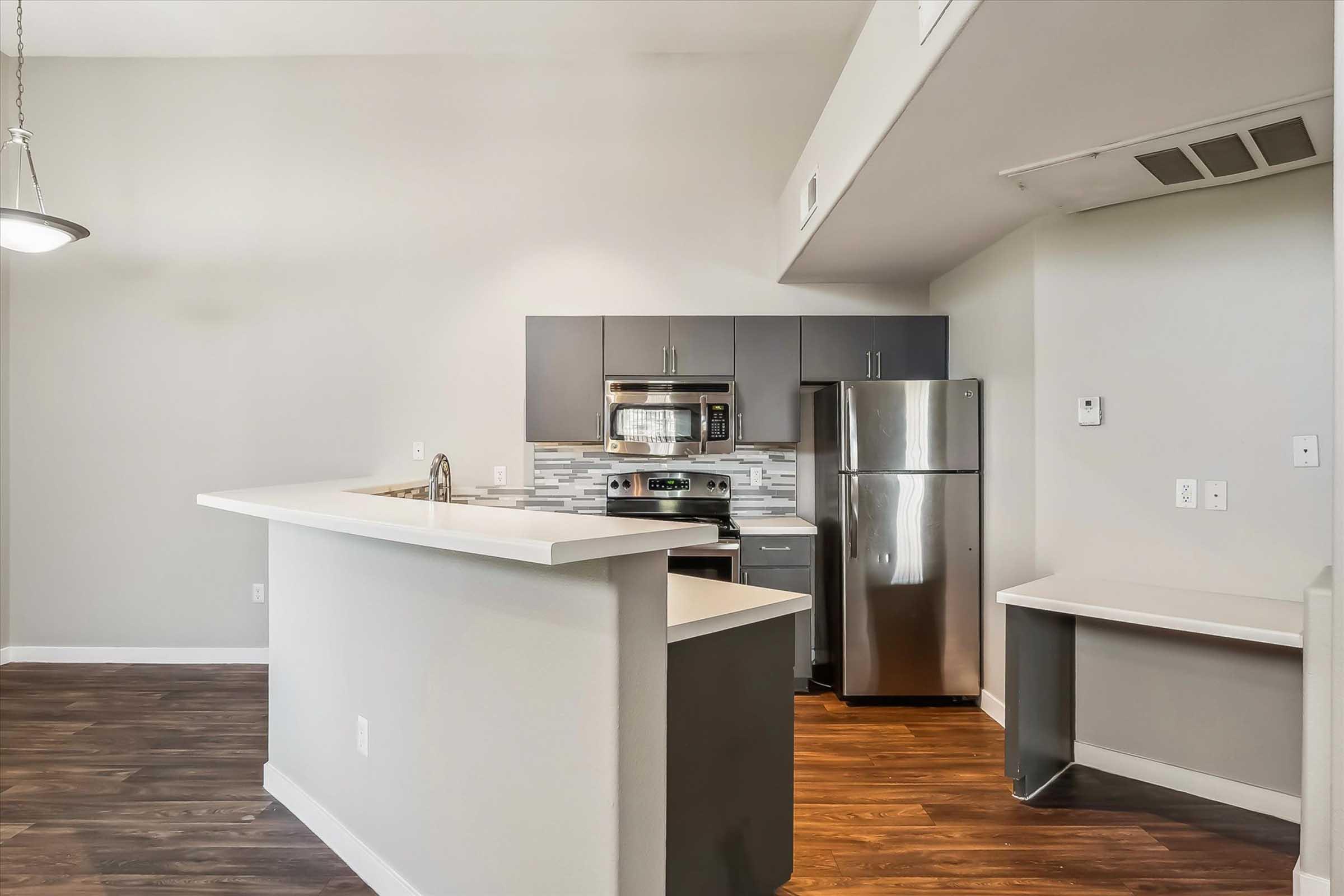 Modern kitchen featuring a gray and white color scheme, with stainless steel appliances, including a microwave and refrigerator. The kitchen island provides a breakfast bar, and there is a small desk area adjacent to the fitted cabinetry. The flooring is a warm wood tone, adding a cozy feel to the space.