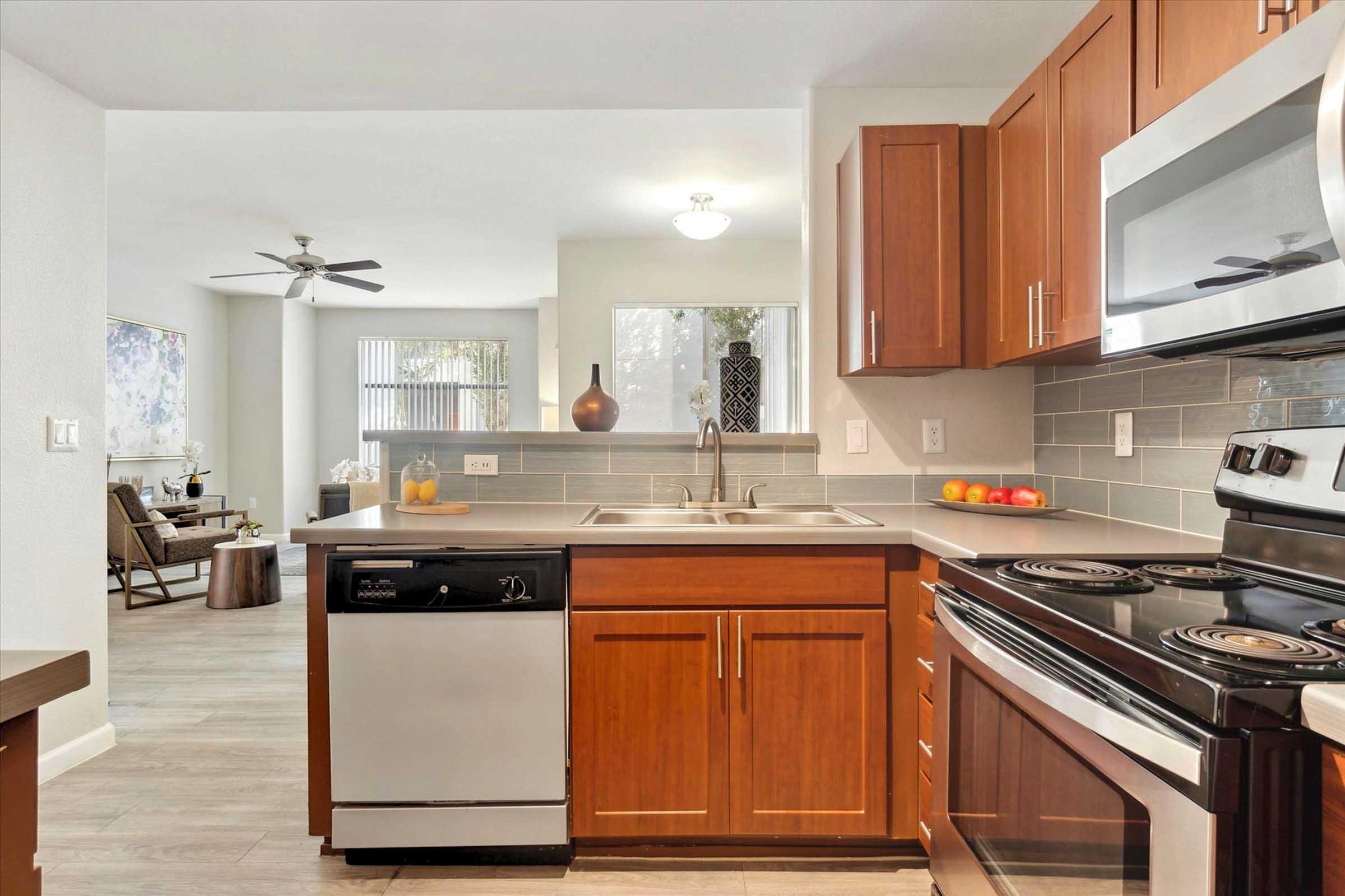 Modern kitchen featuring wooden cabinetry, stainless steel appliances including a dishwasher and oven, and a double sink. The space is well-lit with natural light coming through a window, and includes decorative items like fruits and a vase. There is a living area visible in the background.