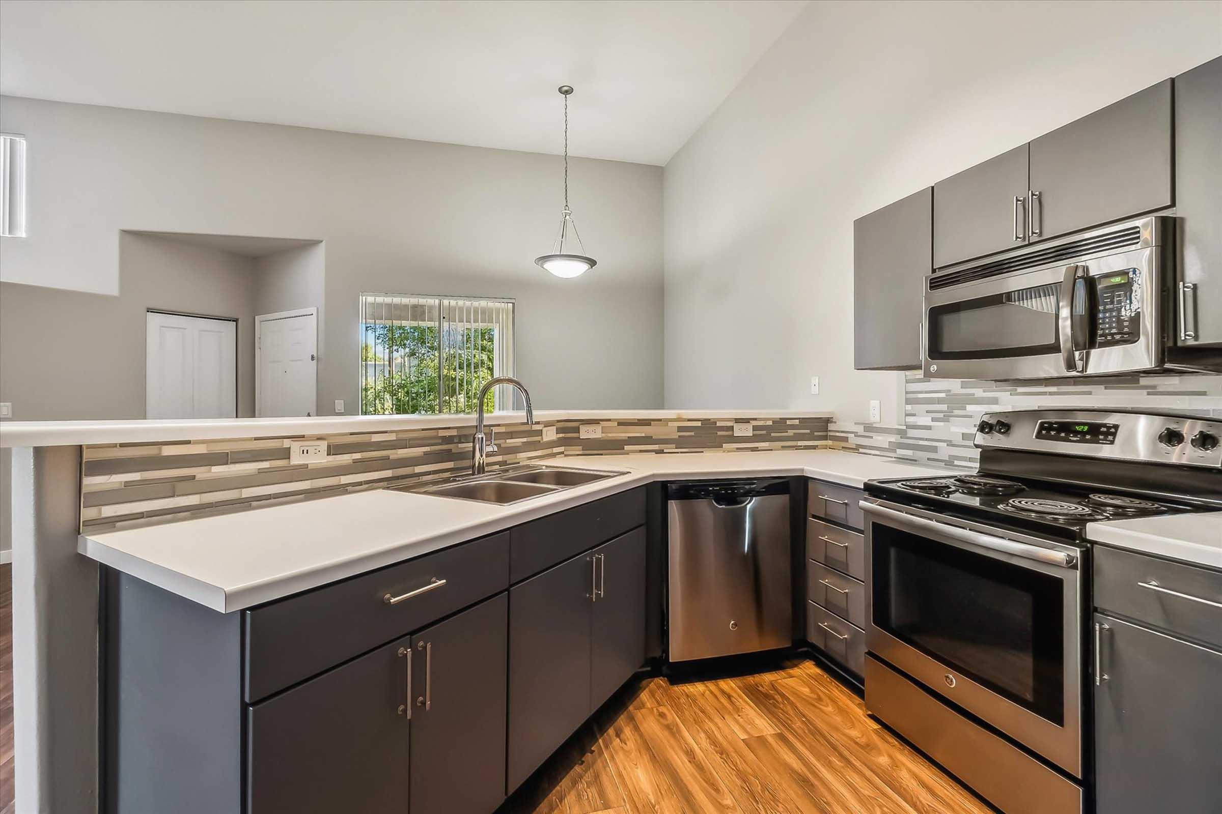 Modern kitchen featuring dark gray cabinetry, stainless steel appliances, and a stylish backsplash. The space includes a sink with a sleek faucet, an electric stove, and a refrigerator, all located in a well-lit area with large windows allowing natural light.