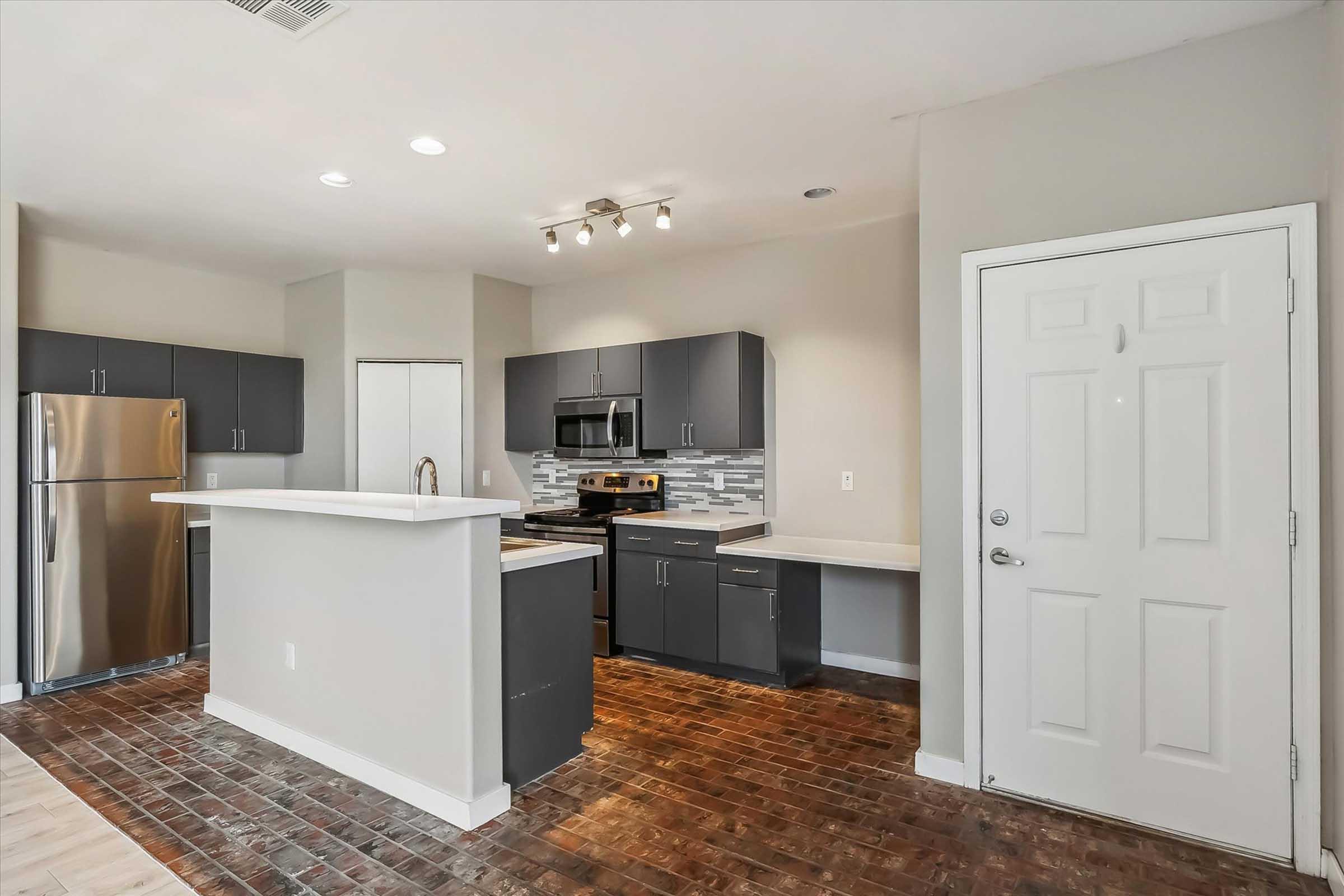 Modern kitchen featuring stainless steel appliances, including a refrigerator and oven. Dark cabinetry complements a light countertop. A gray and white backsplash adds style, while the open layout includes a doorway to another space and warm wood-like flooring. Bright lighting enhances the spacious feel.