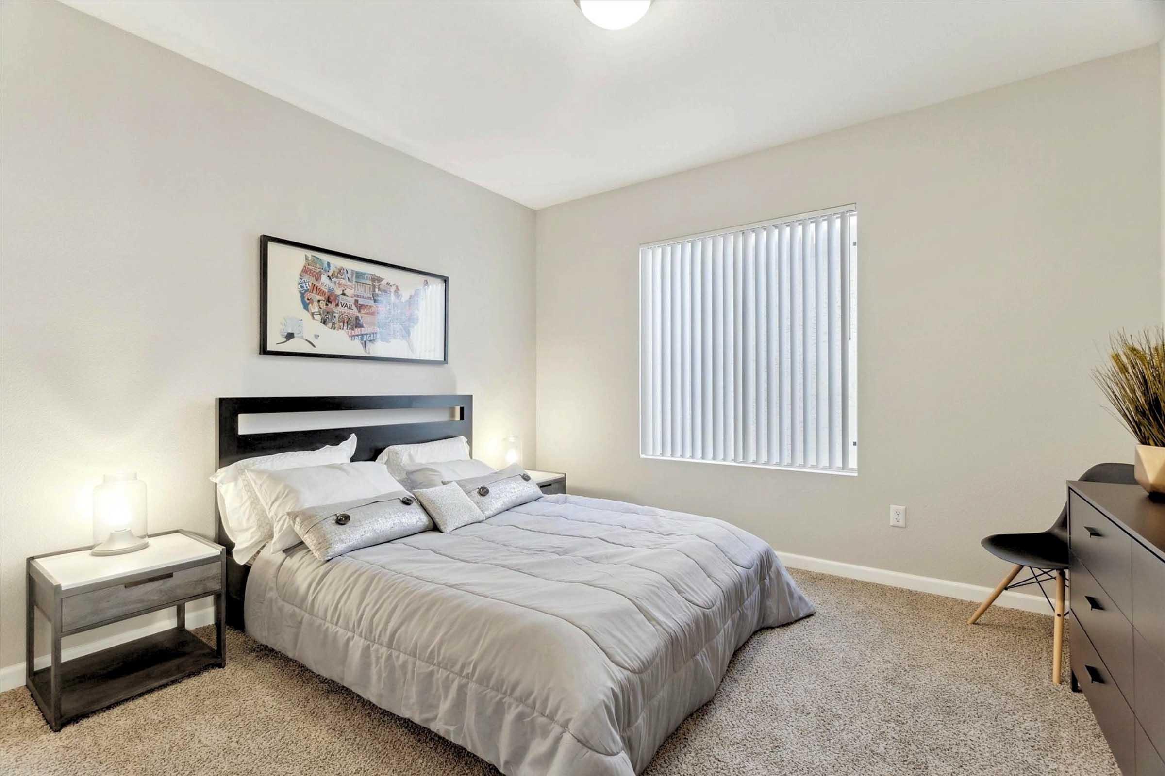A cozy bedroom featuring a queen-sized bed with gray bedding, two decorative pillows, and a modern bedside table with a lamp. There is a large window with vertical blinds allowing natural light in, and minimalistic decor including a framed artwork and a small chair against the wall. The room has neutral tones and carpeted flooring.