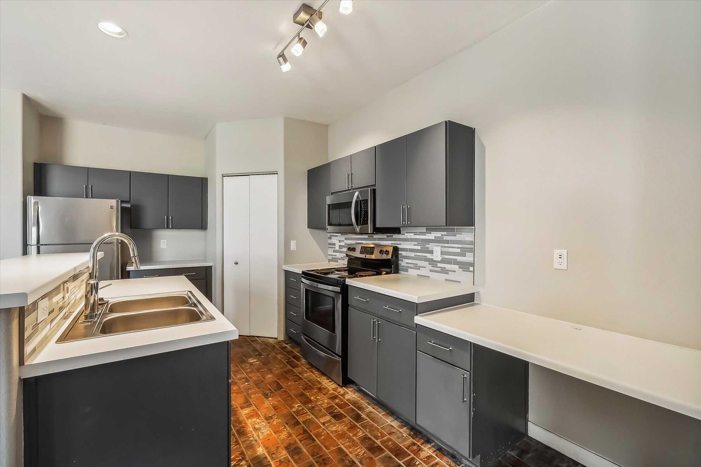 Modern kitchen featuring dark cabinetry, stainless steel appliances, and a tiled backsplash. The spacious layout includes a double sink, a countertop for prep space, and ample storage. Natural light illuminates the room, highlighting the sleek design and warm-toned flooring.
