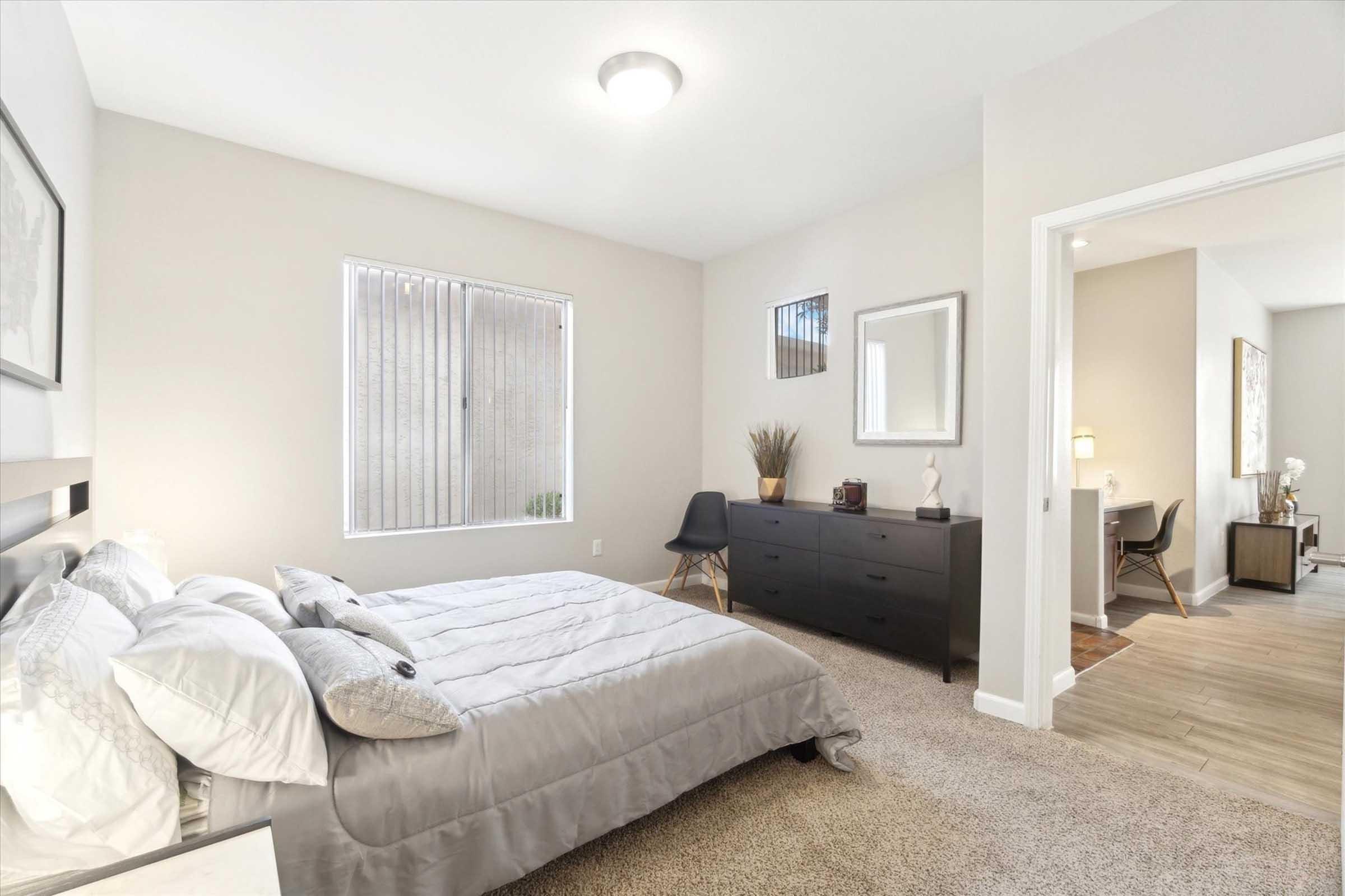 A cozy bedroom featuring a queen-sized bed with light gray bedding, a wooden dresser, and a black chair. Natural light filters through a window with vertical blinds, and there is a mirror on the wall. The room has neutral tones and soft carpeting, creating a warm and inviting atmosphere.