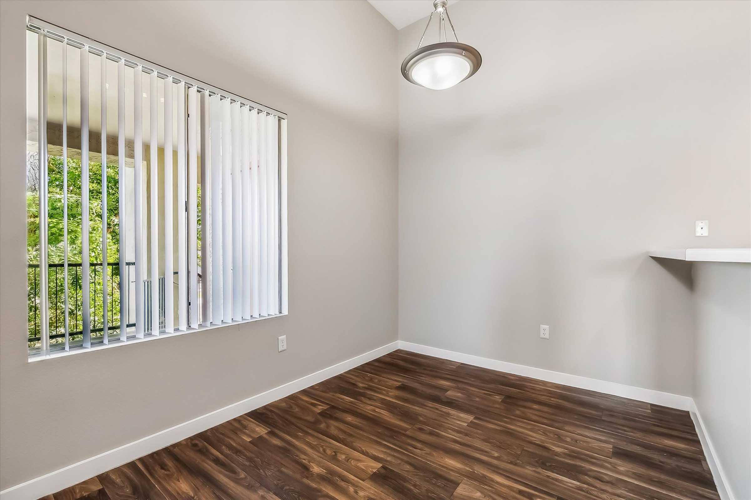 Bright, unfurnished room with wood-like flooring, a large window featuring vertical blinds, and a hanging light fixture. The walls are painted in a neutral color, enhancing the spacious feel. There is a bar counter on one side, creating an inviting layout for potential furniture arrangement.