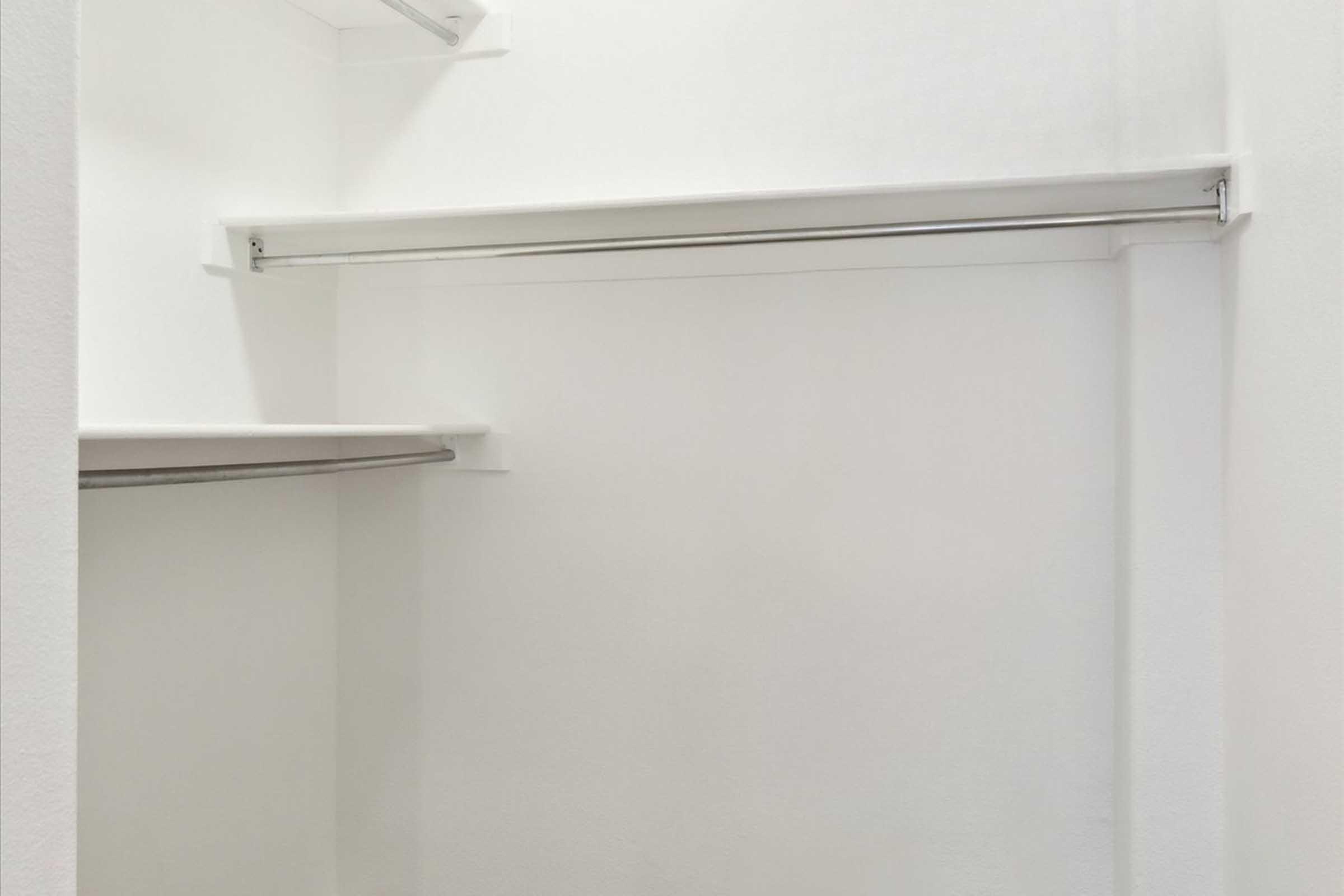 A clean, empty closet with white walls and shelves. There is a horizontal rod for hanging clothes, and the space is well-lit, giving a spacious and minimalist appearance.