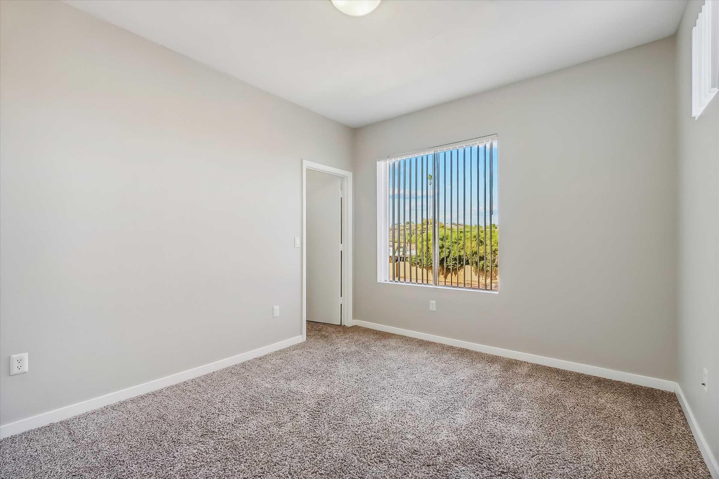 Empty room with light gray walls and carpeted flooring. A window with vertical blinds allows natural light, revealing a view of greenery outside. A door on the left leads to another space. The room is clean and spacious, designed for versatile use.