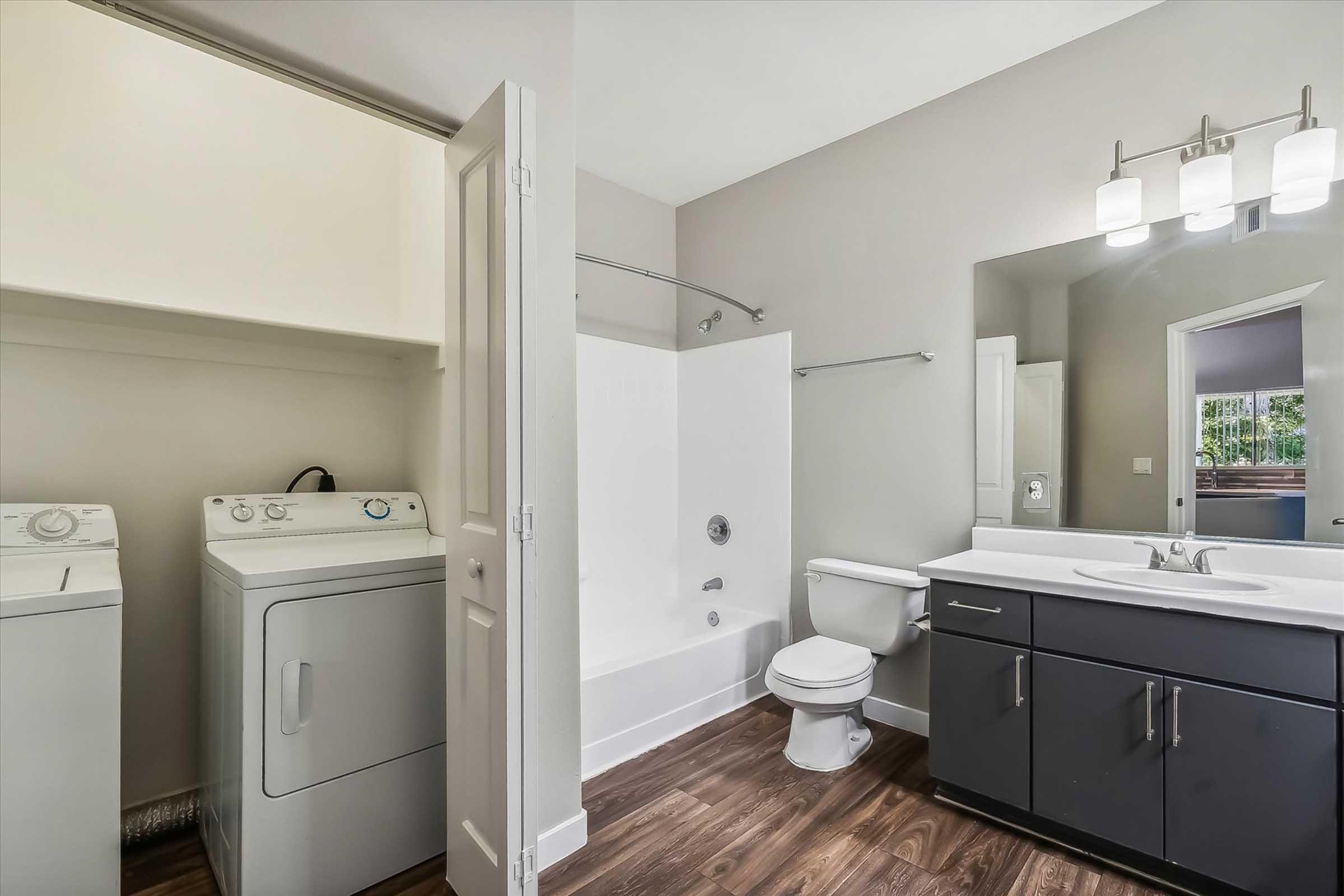 Modern laundry area featuring a stacked washer and dryer beside a bathroom setup that includes a bathtub, toilet, and a sleek vanity with a mirror. The room has light-colored walls and dark wood-like flooring, creating a spacious and clean aesthetic.