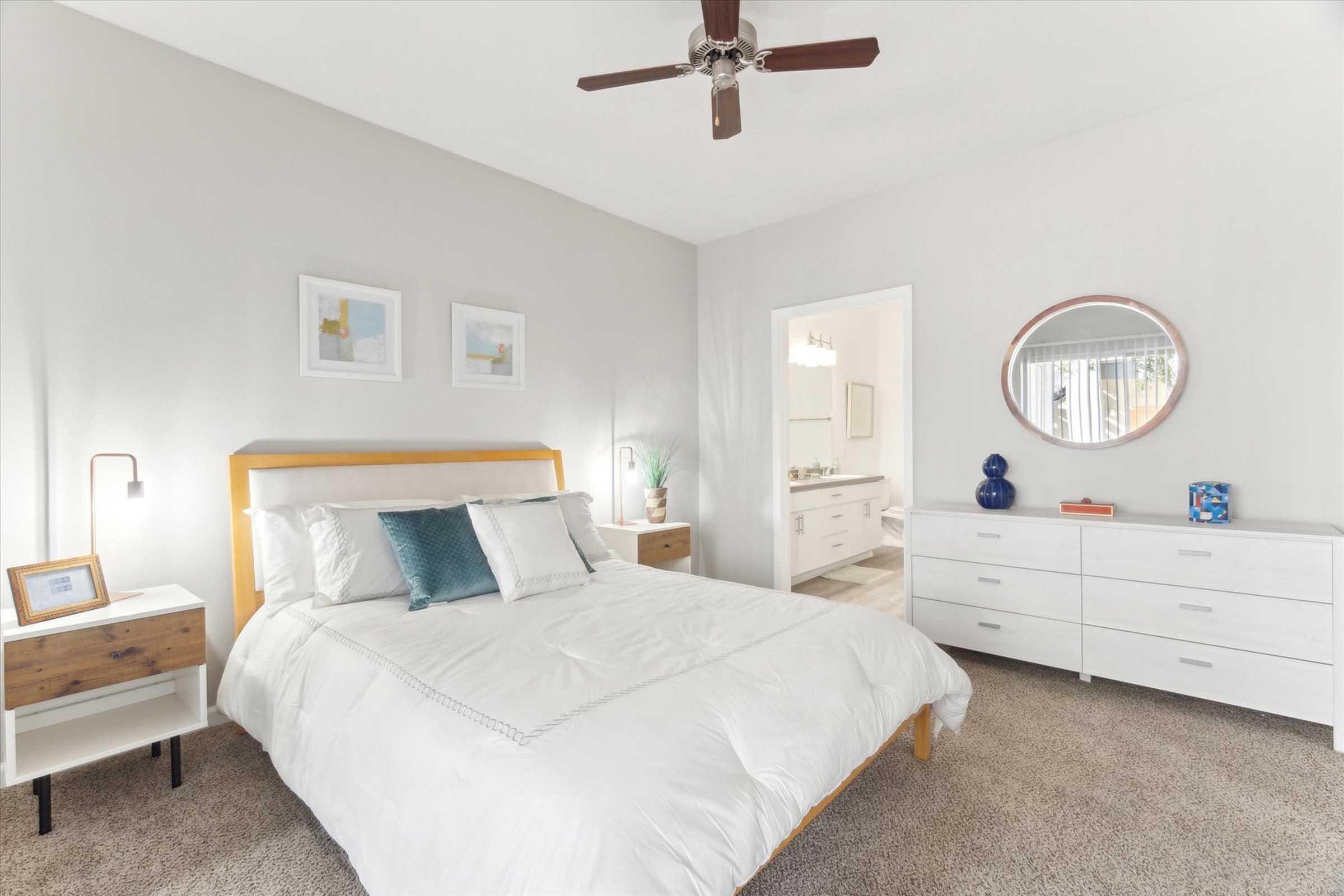 A modern bedroom featuring a white bed with decorative pillows, a wooden headboard, and two bedside tables. A ceiling fan is overhead. The walls are painted light gray, and there are framed pictures and a round mirror on the wall. A doorway leads to an adjacent bathroom, and the floor is carpeted in a neutral tone.