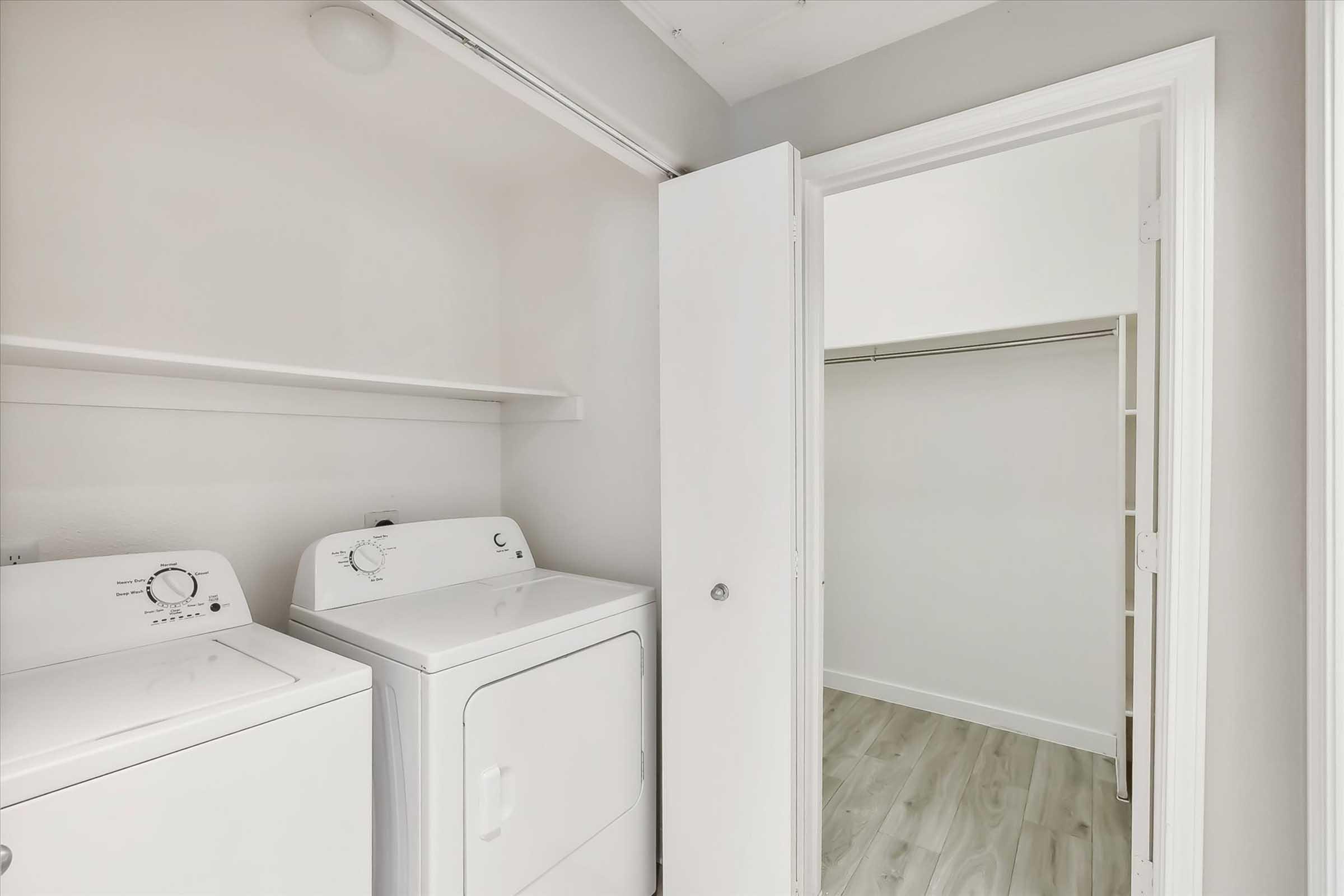 Interior view of a laundry area featuring a stacked washer and dryer. The area is clean and well-lit, with light-colored walls and wood-like flooring. A separate open closet space is visible through an adjacent door.