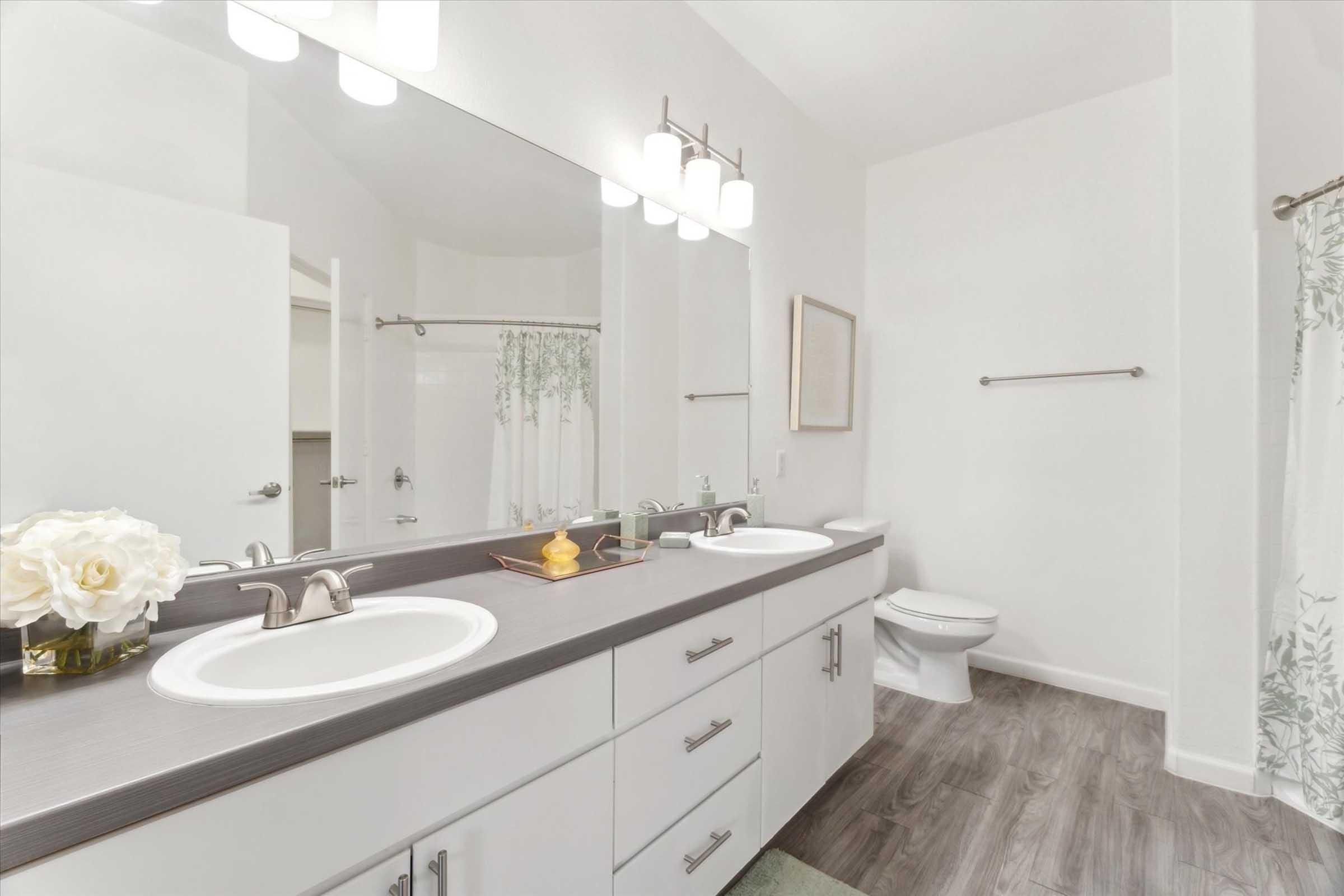A modern bathroom featuring dual sinks with chrome faucets, a large mirror, and a light-colored vanity. There is a white toilet in the background and a green floral shower curtain. A small decorative item with a yellow accent is placed on the countertop, along with a vase of white flowers. The flooring is a light wood texture.