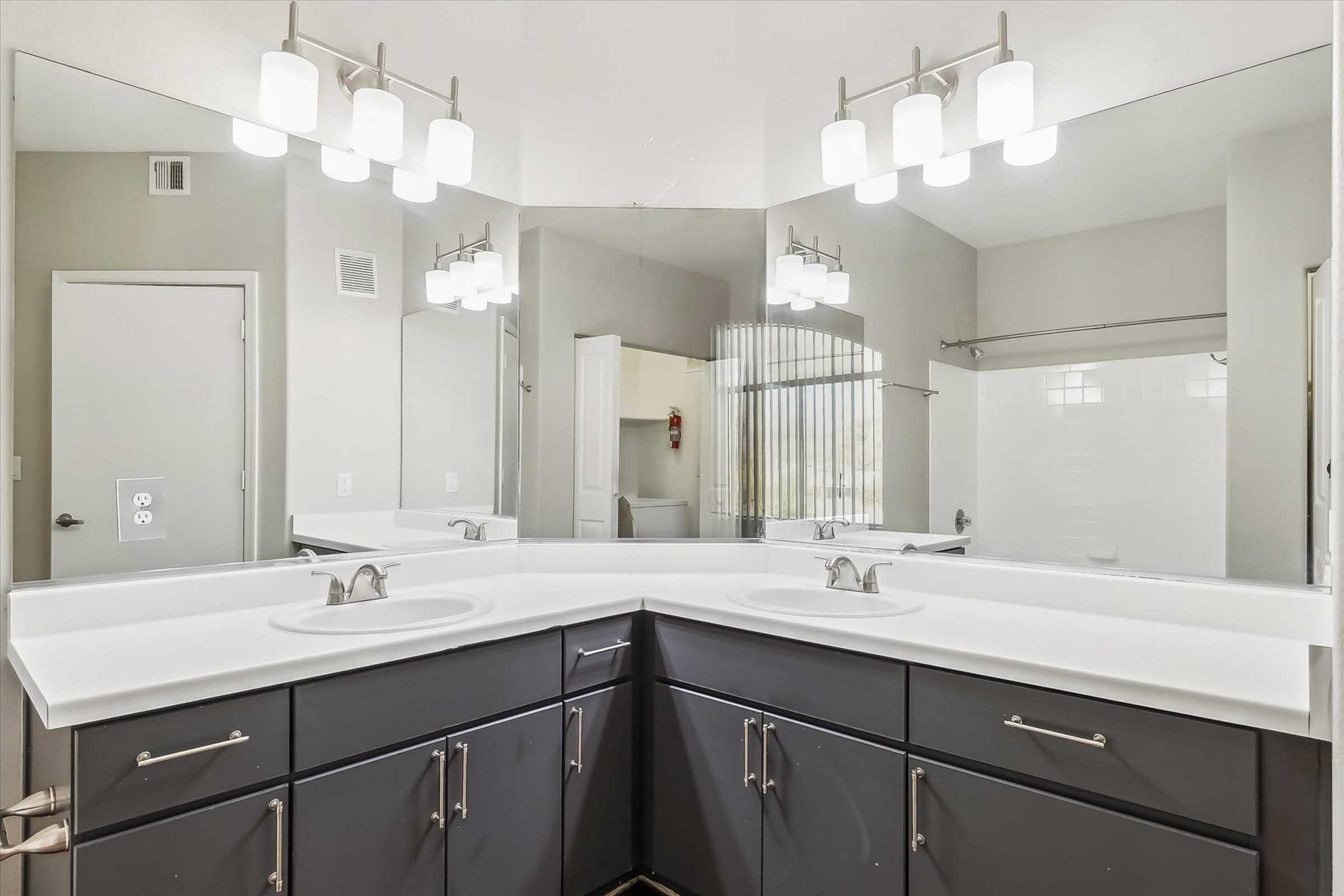 A modern bathroom with a dual sink vanity featuring dark cabinetry and a white countertop. The mirrors above the sinks reflect the space, while stylish light fixtures hang overhead. In the background, a shower area is partially visible, with a beige wall and a glass partition. The overall decor is sleek and contemporary.