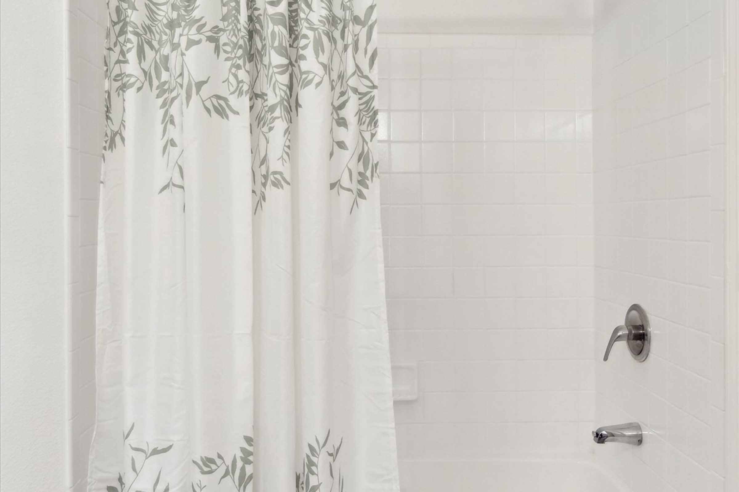 A clean, white bathroom featuring a shower with a light-colored curtain adorned with green leaf patterns. The walls and tiles are white, creating a fresh and airy atmosphere. A silver faucet and showerhead are visible, completing the modern aesthetic.