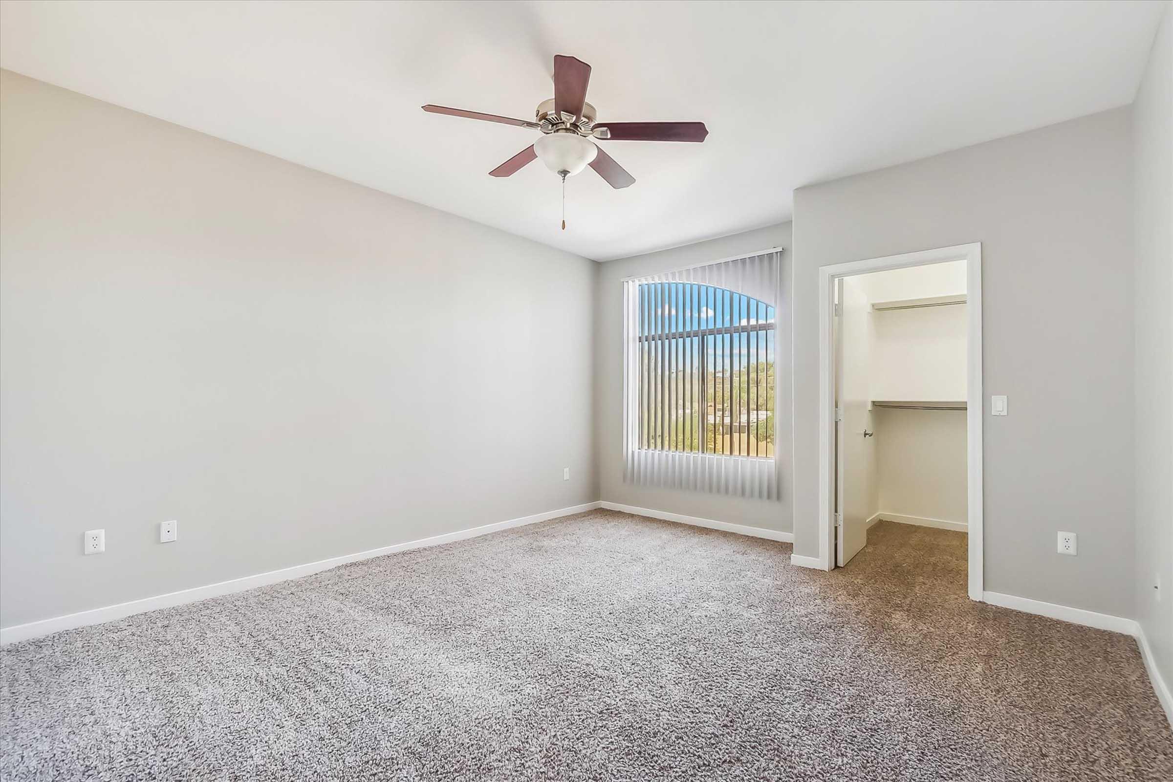 A well-lit, spacious bedroom featuring neutral-colored walls and plush carpet. It includes a ceiling fan, large vertical blinds on a window, and a door leading to a closet area. The room is empty, providing a blank canvas for personal decor.