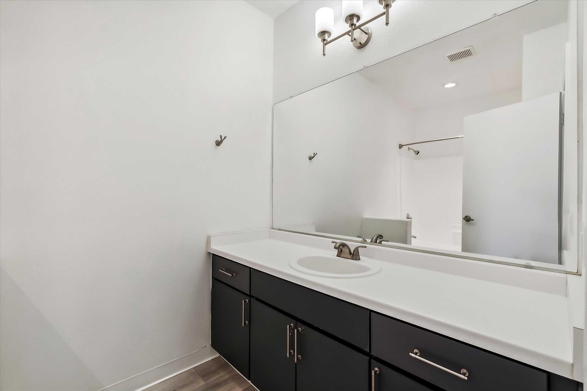 A minimalist bathroom featuring a large mirror above a dark cabinet with a white countertop. Wall-mounted light fixtures illuminate the space. There are hooks on the wall for towels, and a shower area is partially visible in the background. The walls are painted white, giving a clean and spacious feel.