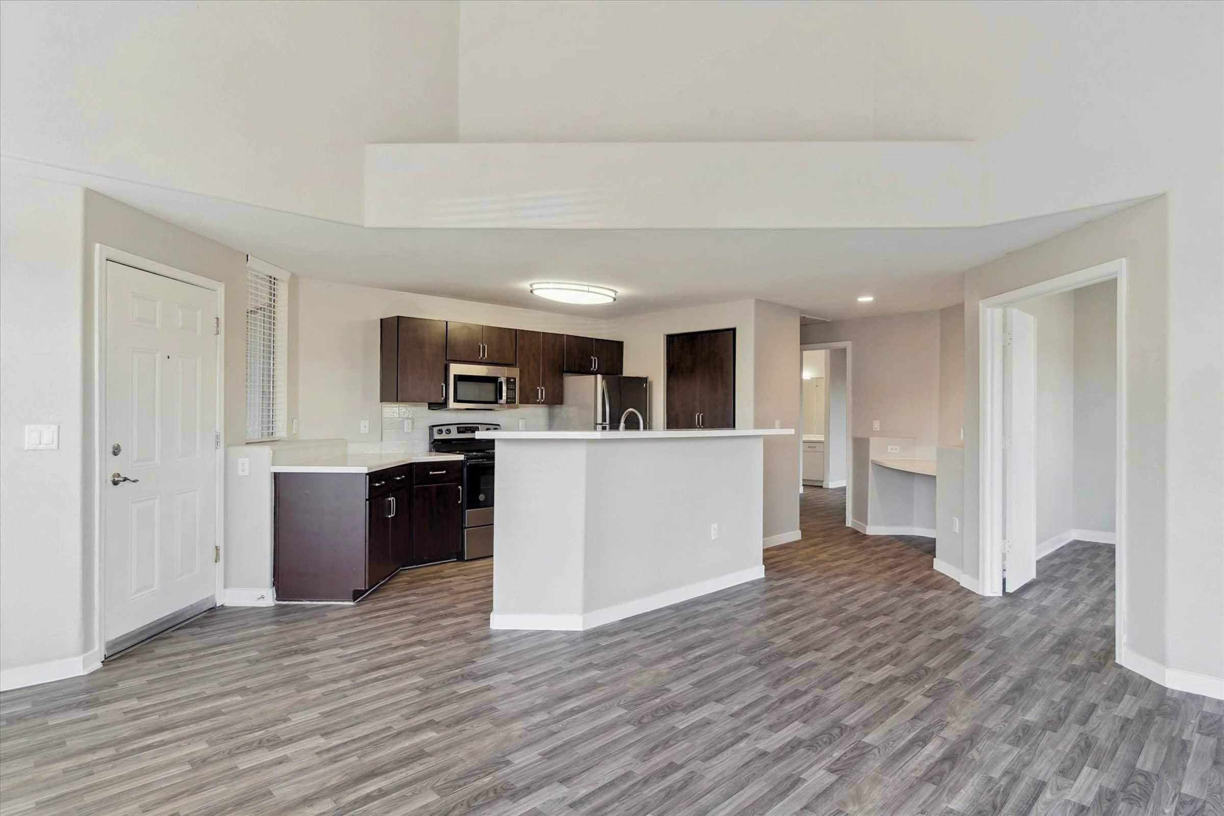 A spacious modern kitchen and living area featuring laminate flooring, dark wood cabinetry, stainless steel appliances, and a large window. The layout includes an open design with a counter separating the kitchen from the living space, creating a bright and inviting atmosphere.