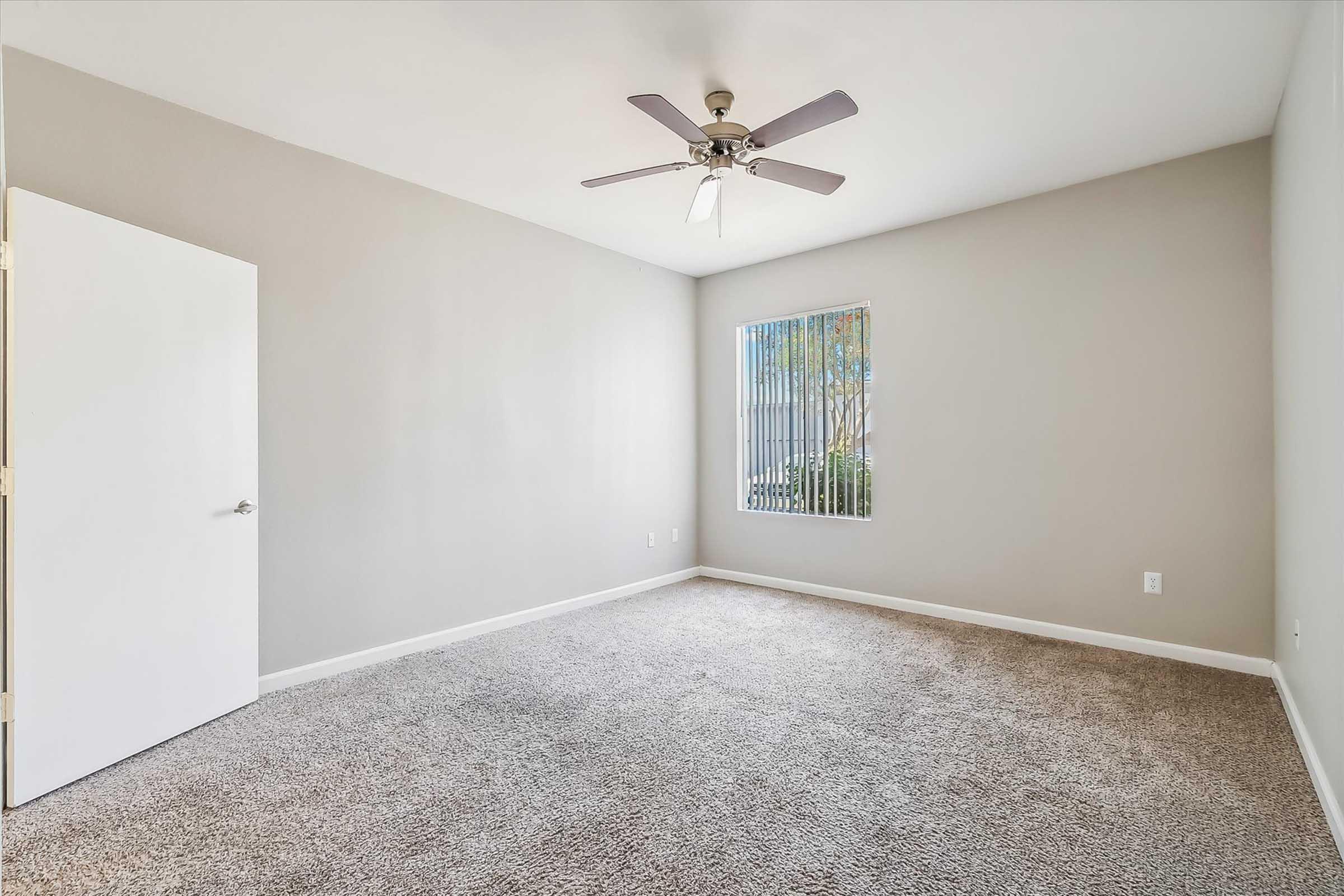 Empty room with light gray walls and beige carpet. A ceiling fan is mounted in the center, and there is a single window with vertical blinds allowing natural light to enter. A white door on the left leads to another space. The room has a spacious and airy feel.