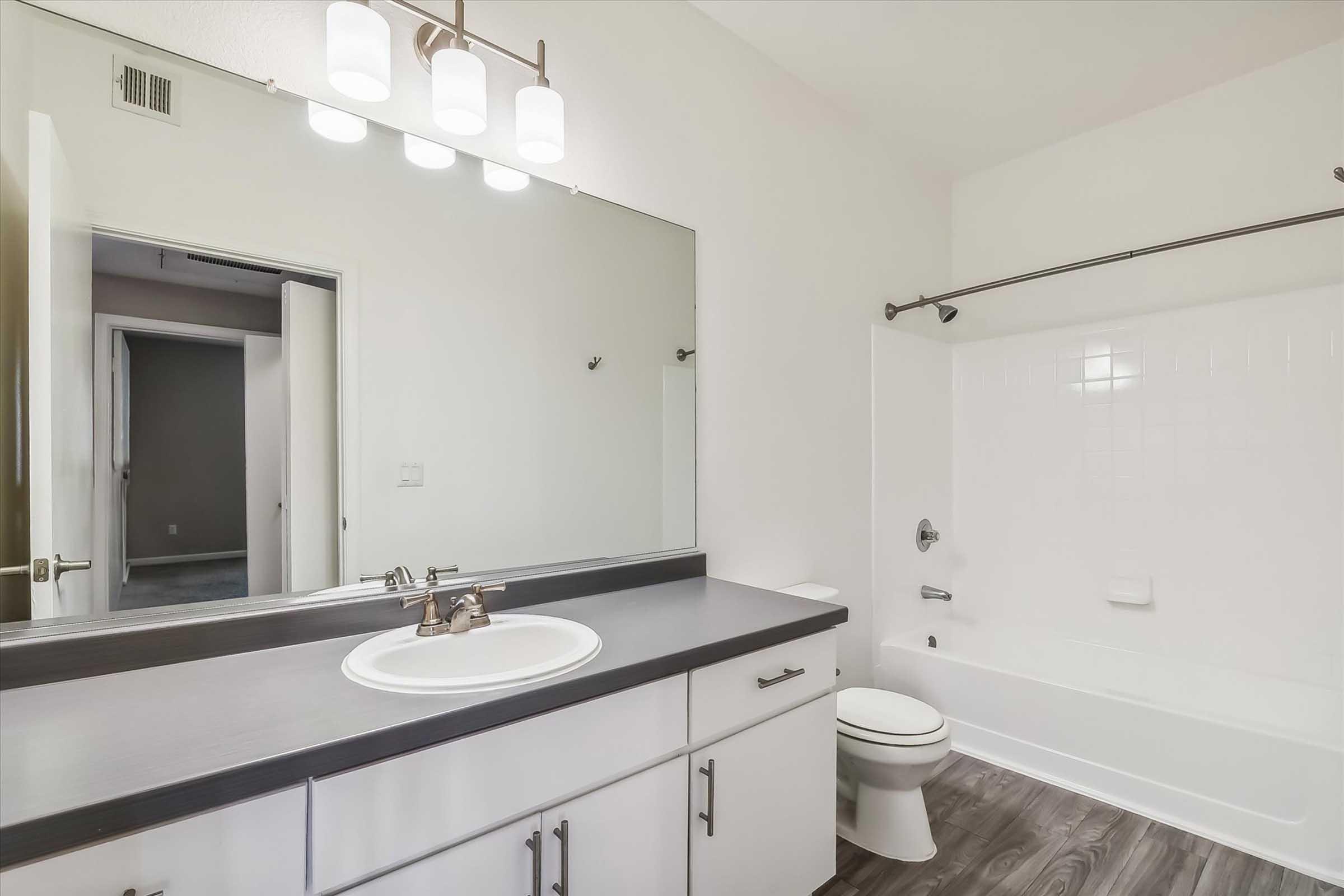 A modern bathroom featuring a large mirror, a white sink, and a countertop with dark gray finishes. The space includes a white bathtub with a shower, a toilet, and a door leading to another room. Soft lighting is provided by overhead fixtures, and the flooring is a wood-like texture.