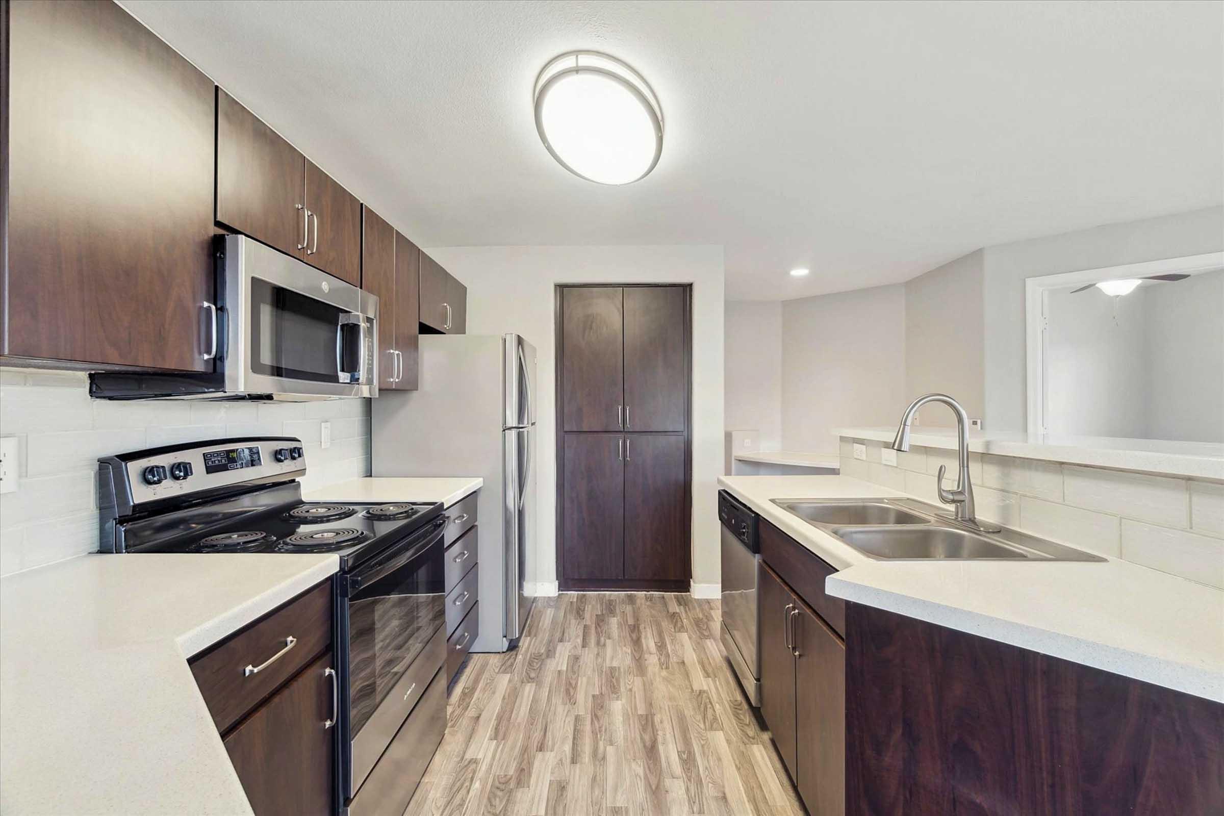 Modern kitchen featuring dark wood cabinets, stainless steel appliances including a microwave, oven, and dishwasher. Light countertops contrast with the cabinetry. The space has a clean, open layout with a large window for natural light and a sleek, contemporary design.