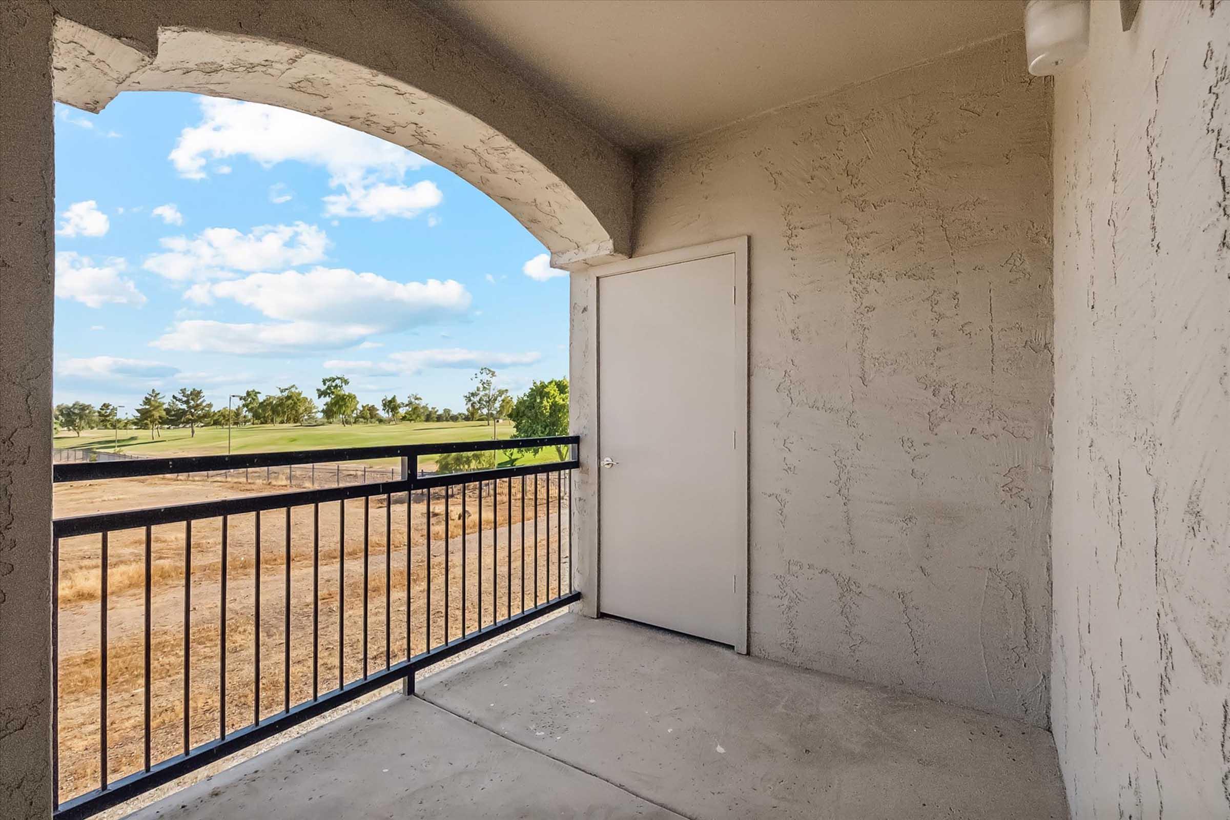 Balcony view featuring a spacious porch with a railing, an open door leading to the interior, and scenic outdoor landscape including trees and open fields against a blue sky with scattered clouds.