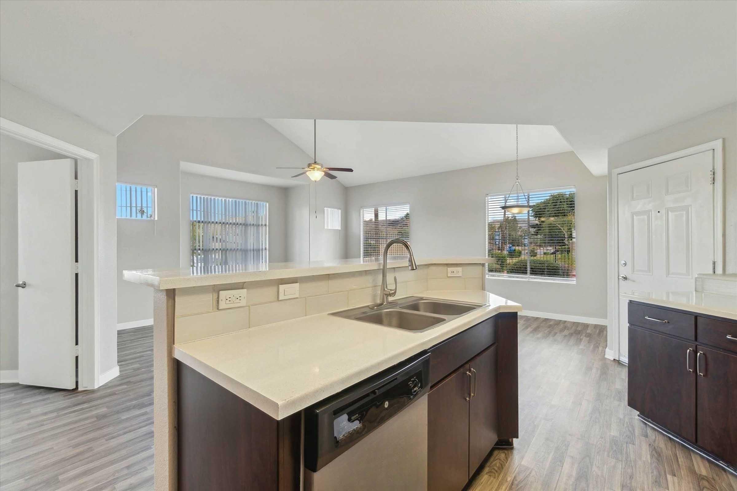 A modern kitchen featuring dark wood cabinets, a stainless steel sink, and a dishwasher. The open layout includes a spacious living area with large windows providing natural light and a view of the outdoors. The walls are painted in a light color, enhancing the bright and airy atmosphere.