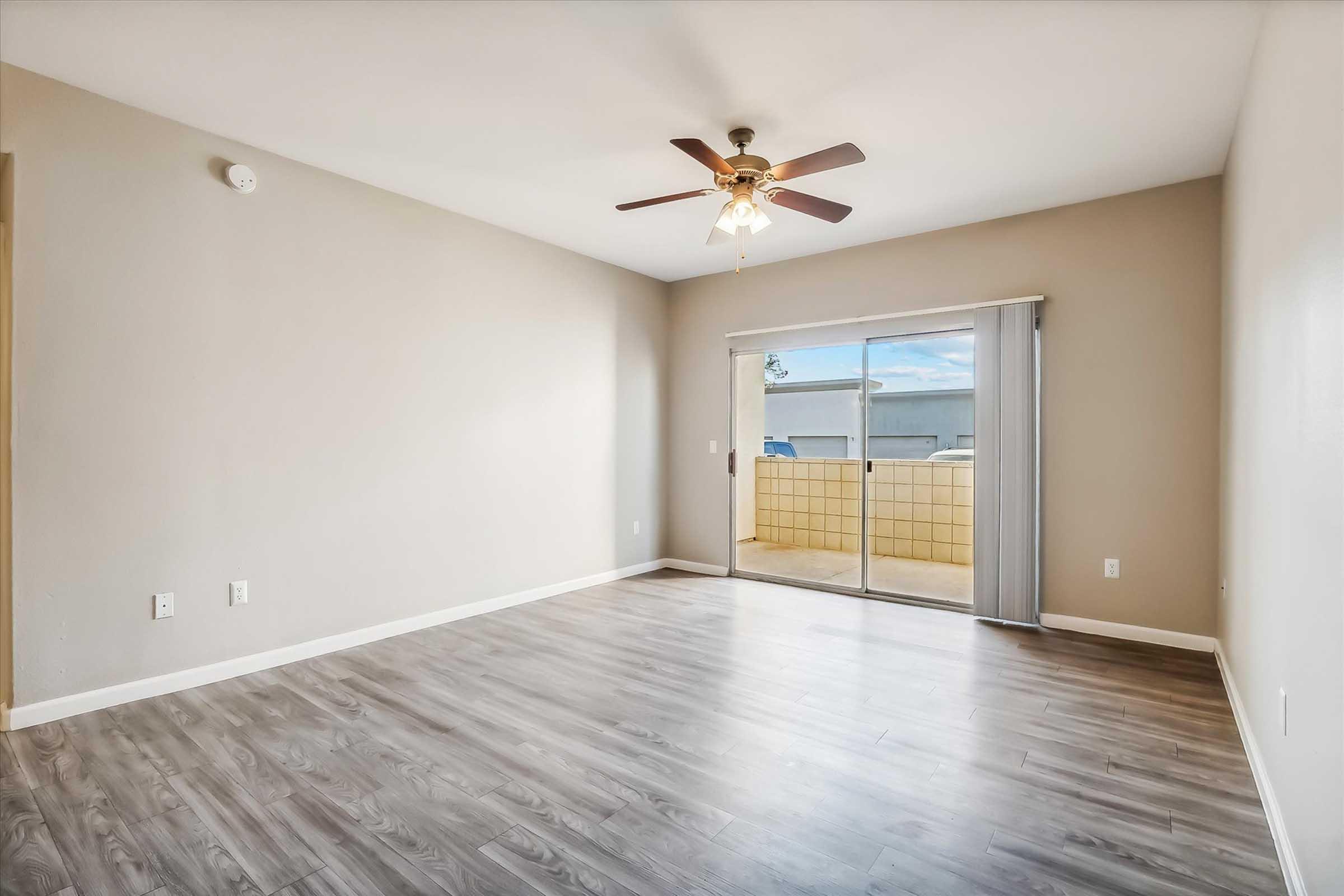 Spacious indoor living area with light brown walls, hardwood flooring, a ceiling fan with wooden blades, and natural light coming through a sliding glass door that leads to a small outdoor patio.