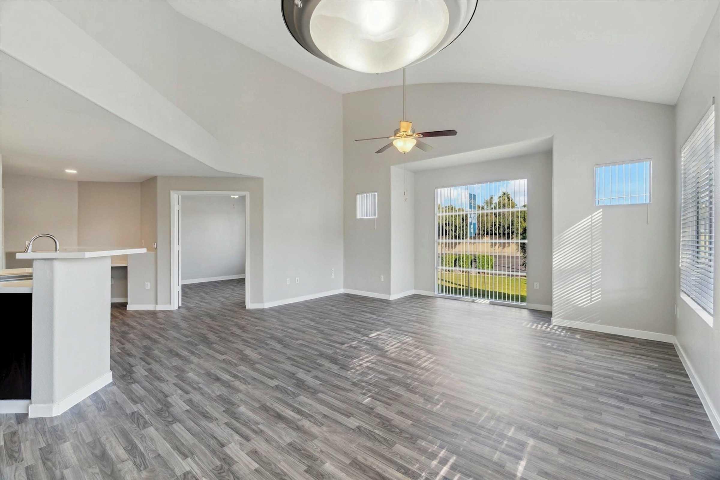 Spacious interior of a modern living area featuring light gray walls, hardwood-style flooring, and large windows letting in natural light. A ceiling fan hangs from a vaulted ceiling, and there is an open layout connecting the living room to the kitchen. A sliding door leads to an outdoor area.