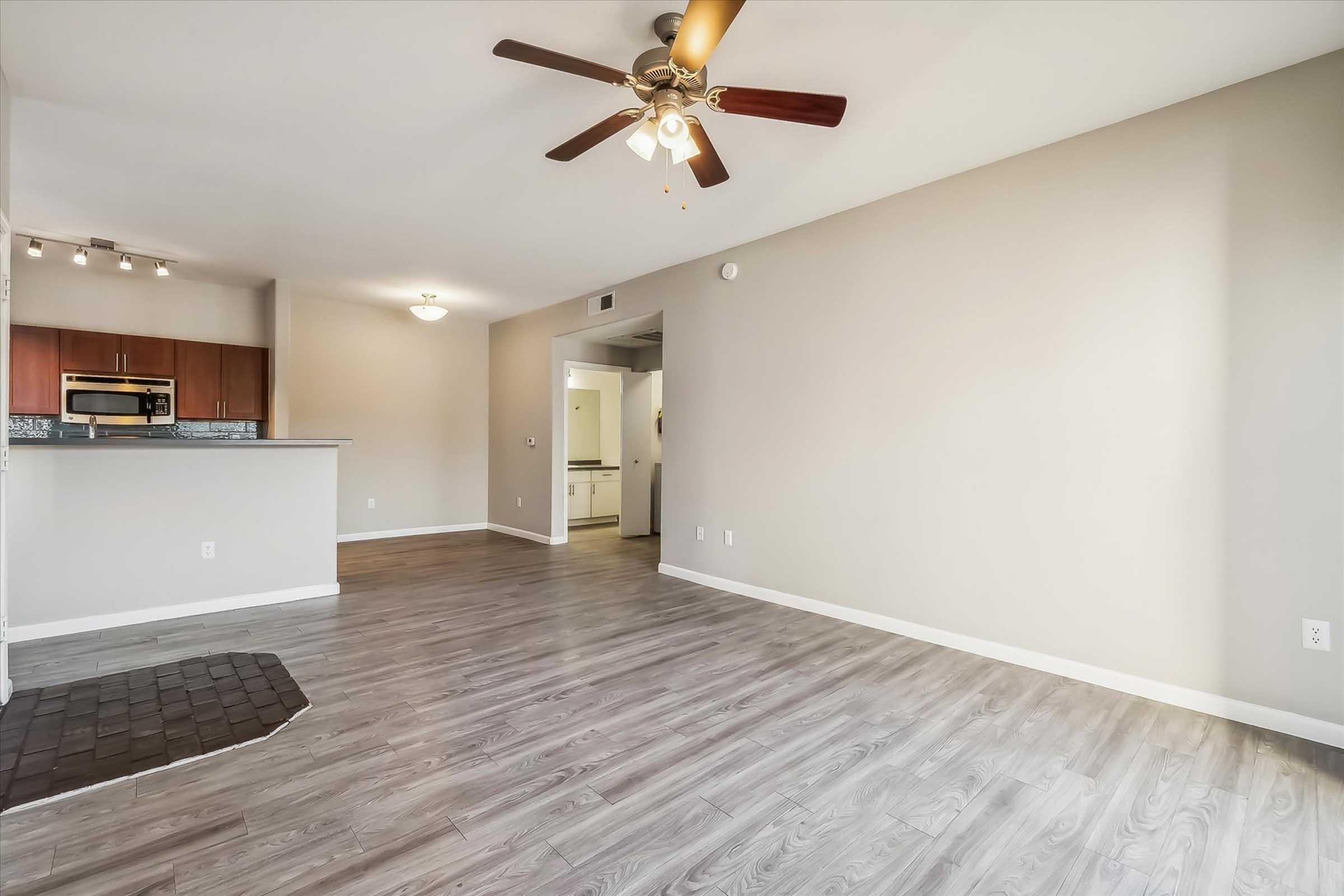 A spacious, well-lit living area with light gray walls and hardwood flooring. A ceiling fan with wooden blades hangs from the ceiling. To the left, there is a kitchen area with wooden cabinets and stainless steel appliances. The room is empty, suggesting it's ready for furniture or decoration.