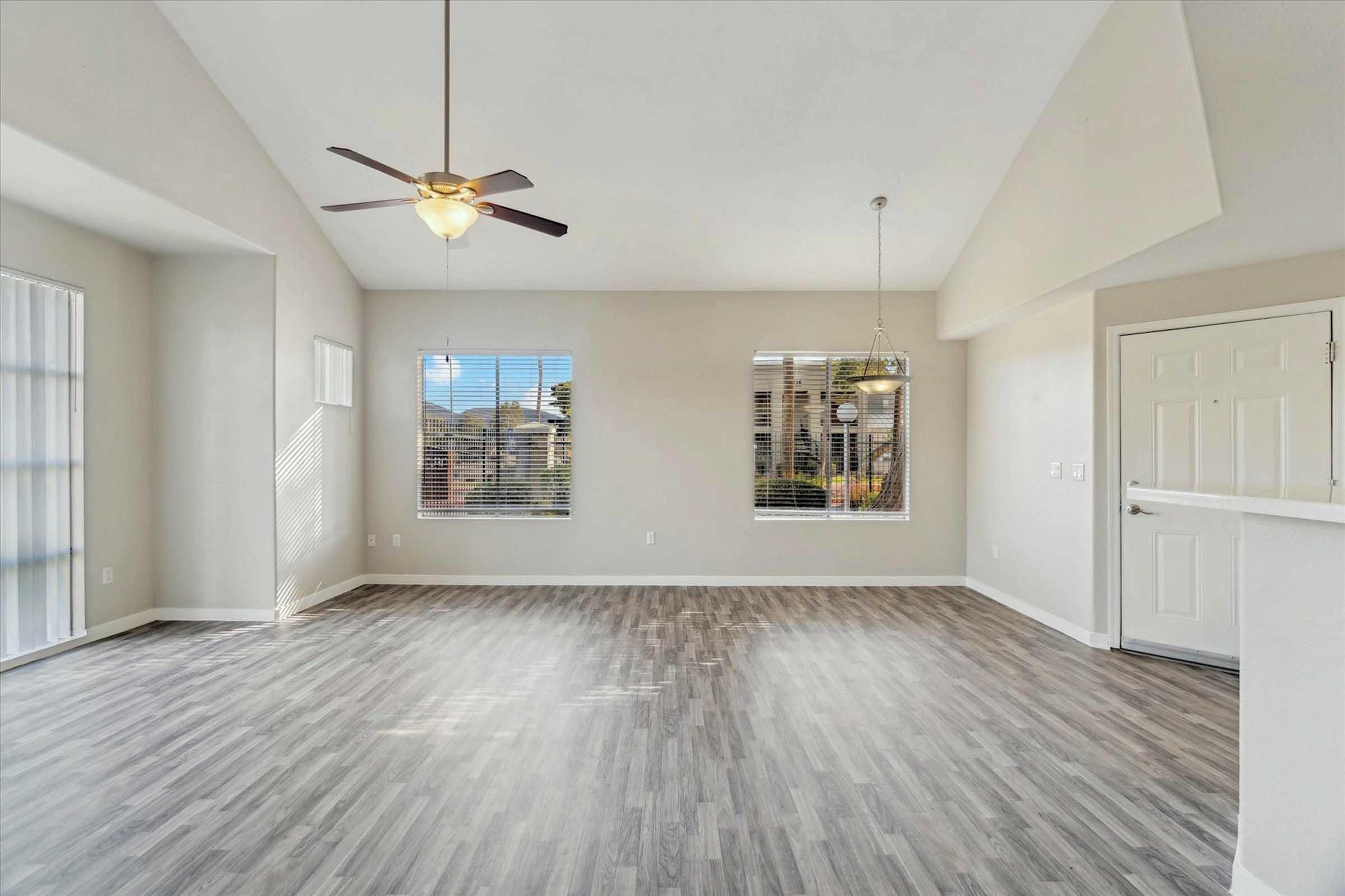 A spacious, well-lit living area with light gray wood-like flooring. Features include a ceiling fan, large windows with blinds, and a sliding glass door leading to an outdoor space. The walls are painted a neutral color, and there is a front door visible on the right.